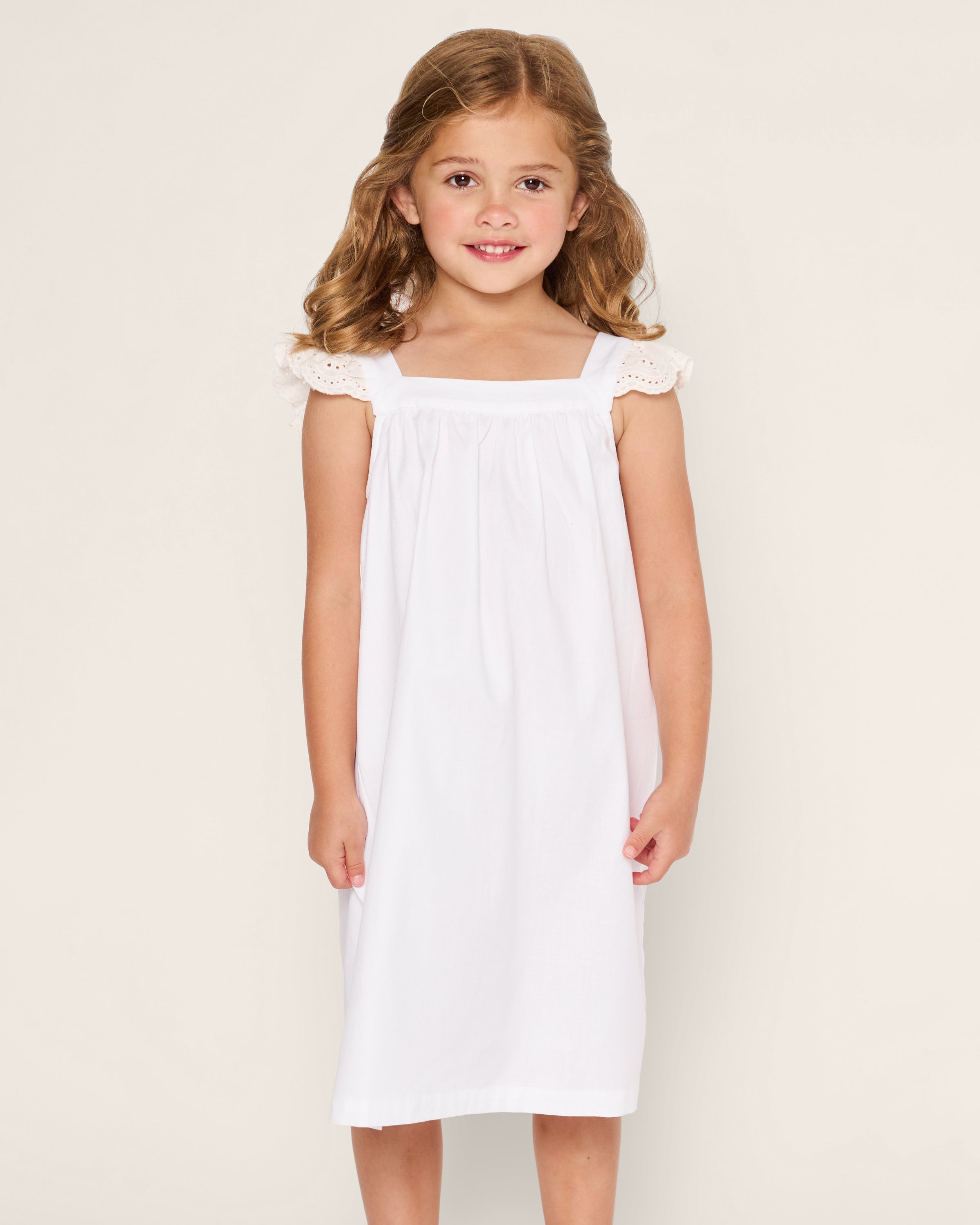 A young girl with long, wavy brown hair smiles wearing Petite Plumes luxurious white Girls Twill Clara Nightgown, featuring lace shoulder details, against a plain light-colored background.