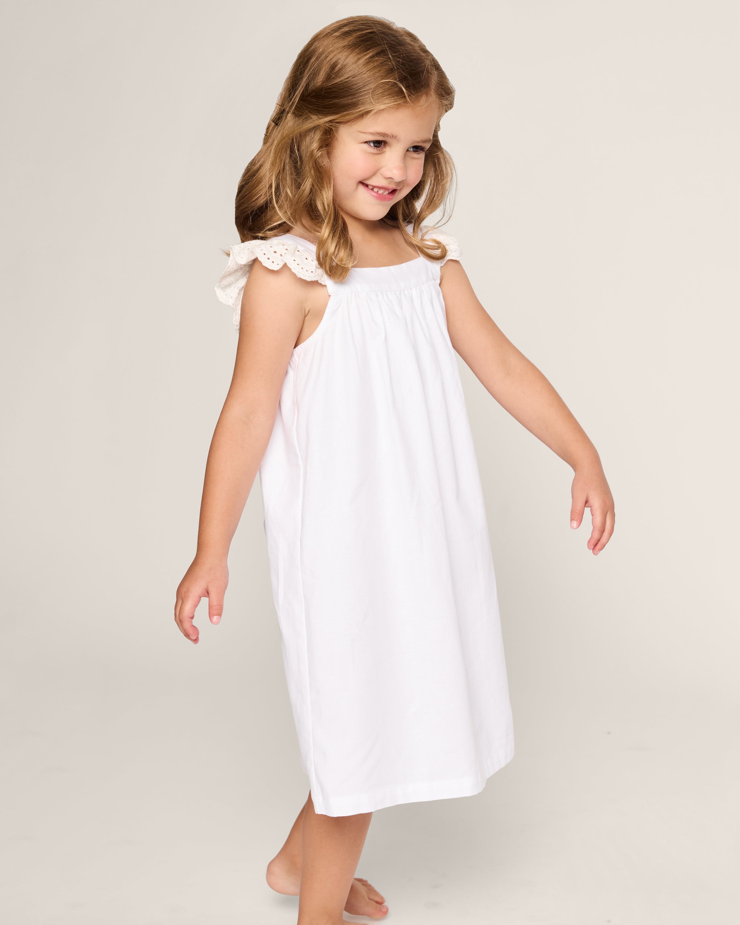 A young girl with brown hair wears the Girls Twill Clara Nightgown in White by Petite Plume. The luxurious cotton gown has lace shoulder straps. She smiles against a plain backdrop, barefoot, with arms slightly extended to the sides.