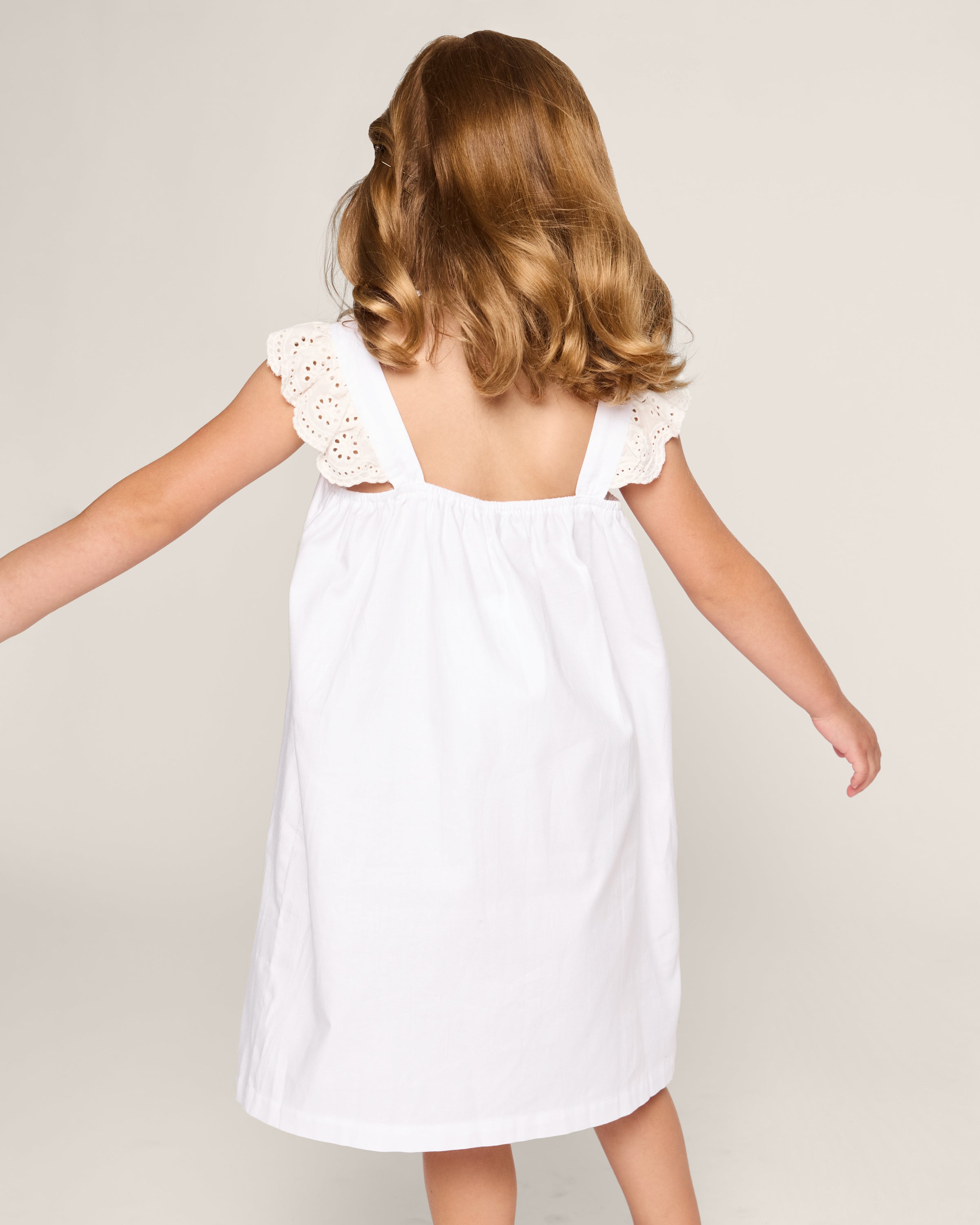 A young girl with wavy, light brown hair faces away in the Petite Plume Girls Twill Clara Nightgown in White. The nightgown features eyelet lace trim on the shoulders. She stands against a plain, light background, arms slightly extended, exuding an aura of airy elegance.