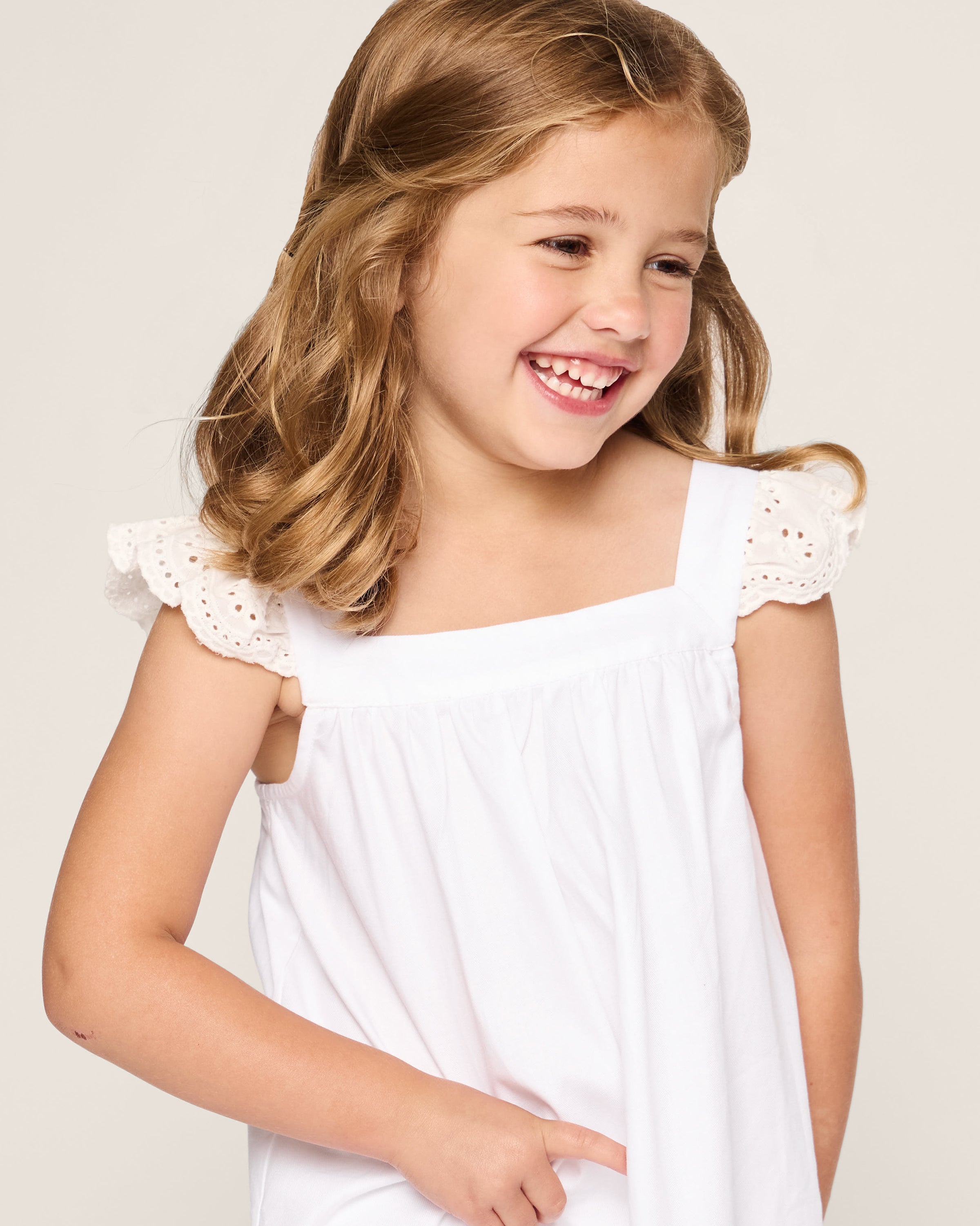A young girl with light brown hair smiles and looks off to the side, wearing the Petite Plume Girls Twill Clara Nightgown in White with eyelet lace cap sleeves, made from luxurious cotton against a plain, light-colored background.