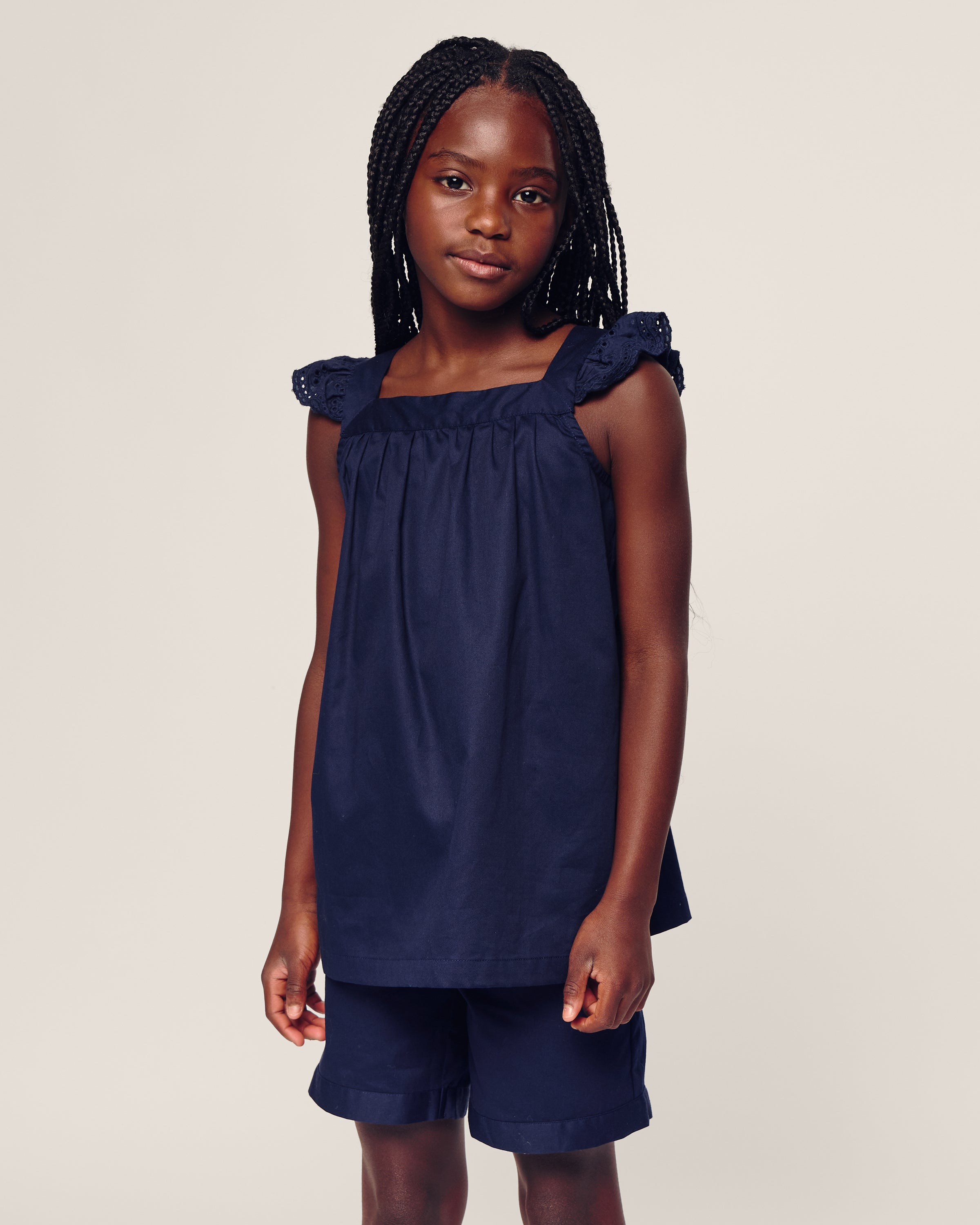 A child with long braided hair wears the Girls Twill Clara Short Set in Navy by Petite Plume, featuring ruffled straps and made of the finest quality cotton. They stand against a plain, light-colored background.