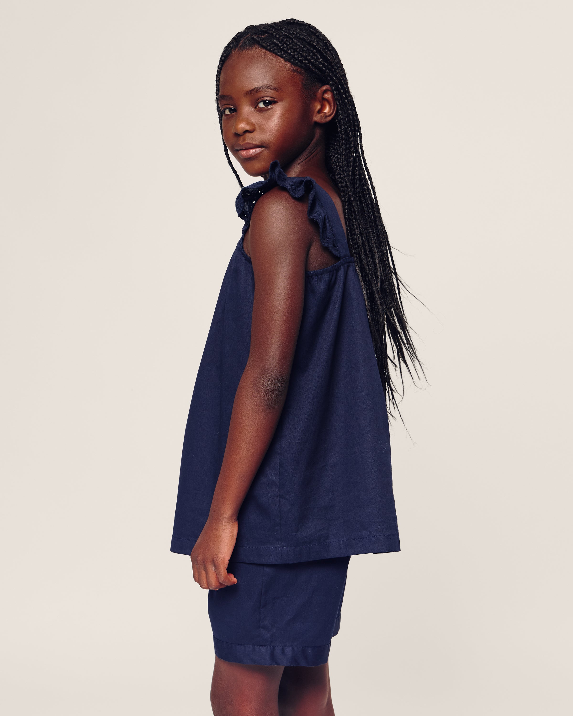 A young girl with long braided hair stands sideways, wearing Petite Plumes Girls Twill Clara Short Set in Navy. Her sleeveless outfit complements the neutral-toned backdrop as she looks at the camera.