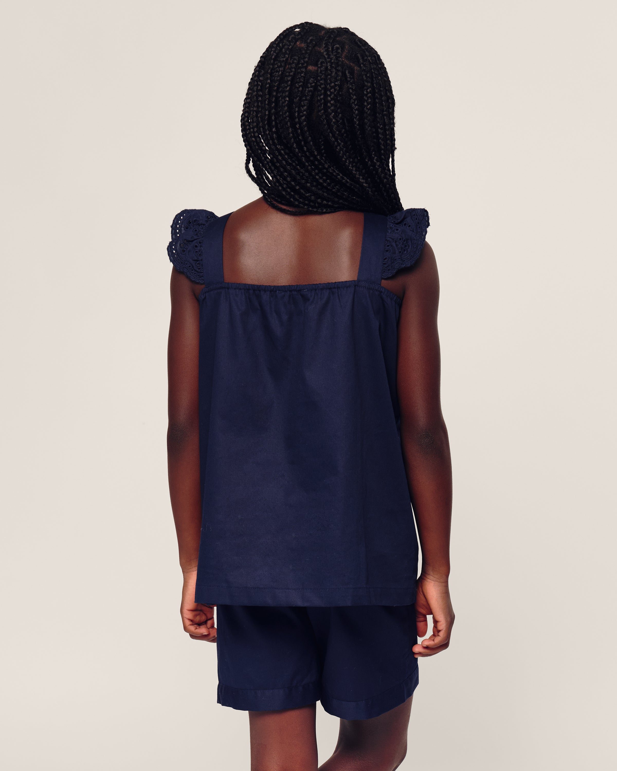 A child with braided hair is seen from behind wearing Petite Plumes Girls Twill Clara Short Set in Navy, showcasing lace details on the shoulders against a plain background.
