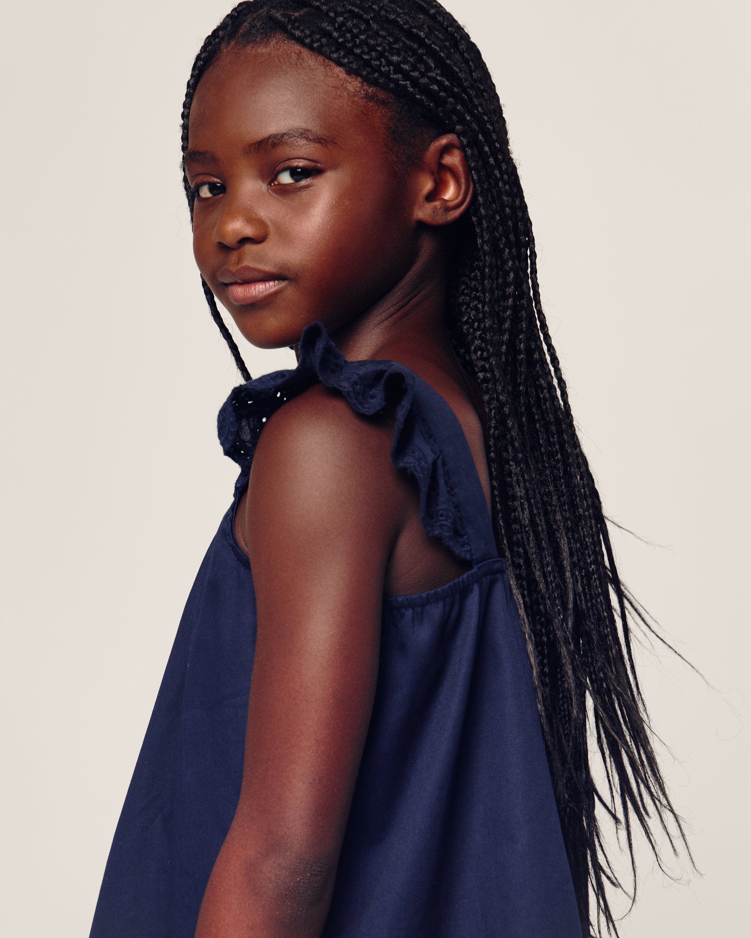 A young girl with long braided hair looks over her shoulder, wearing the Girls Twill Clara Short Set in Navy from Petite Plume. She stands against a plain white background.
