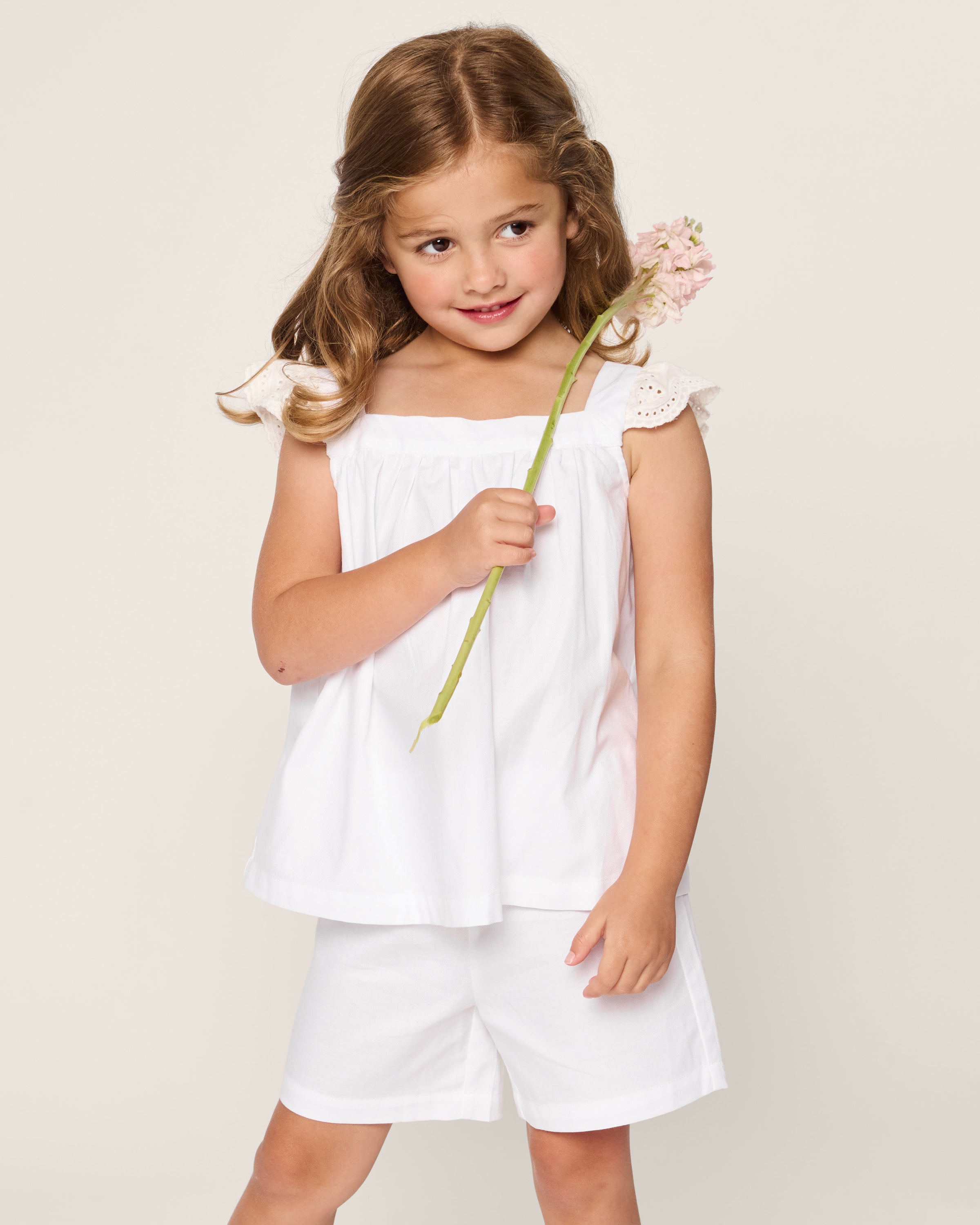 A young girl with light brown hair wears Petite Plumes Girls Twill Clara Short Set in White, holding a pink flower. She stands against a plain background, smiling slightly and looking to the side.