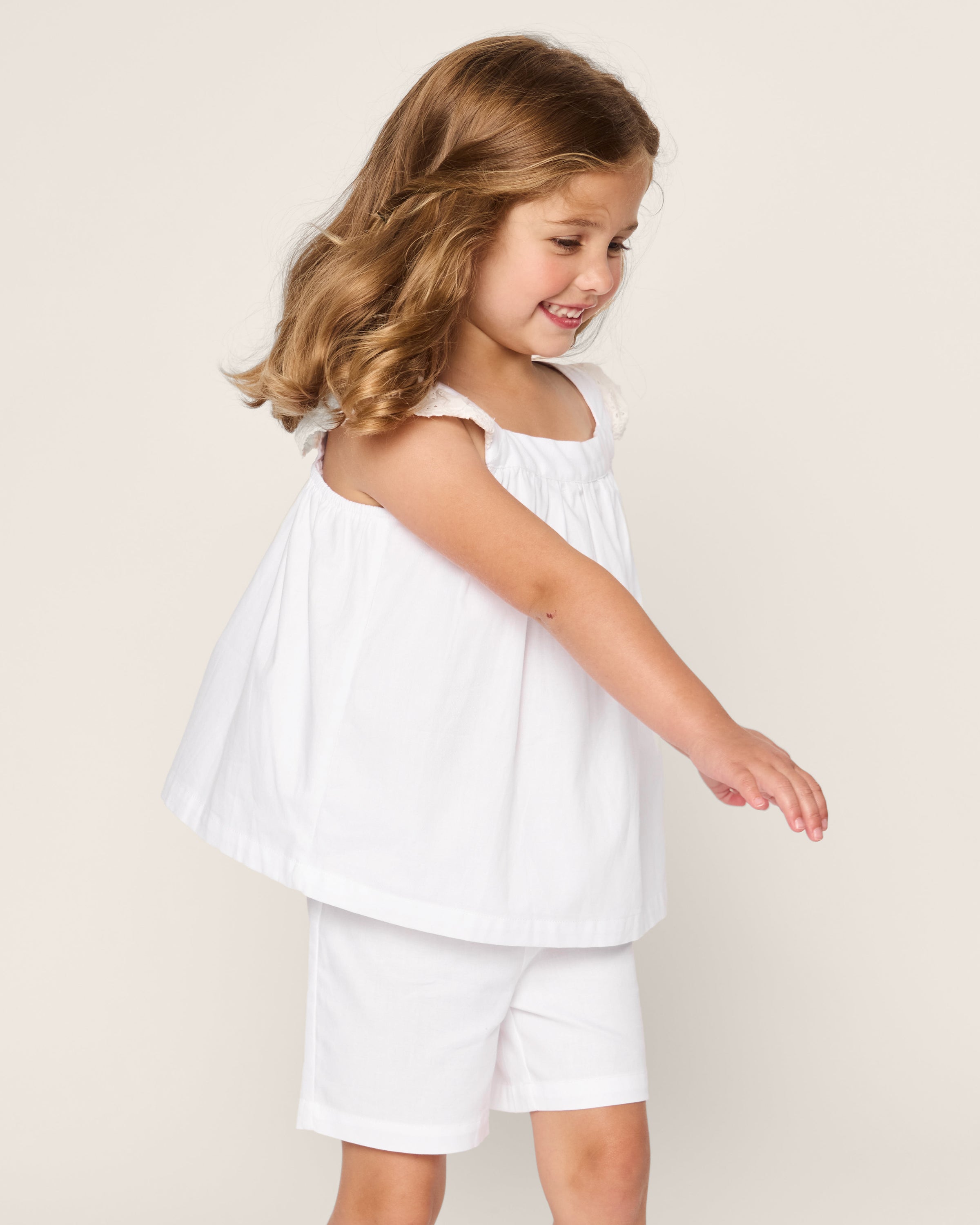 A young girl with wavy brown hair smiles while looking to the side, wearing Petite Plumes Girls Twill Clara Short Set in White. The breezy outfit features a sleeveless white top with ruffled sleeves and matching shorts made of high-quality cotton, set against a plain off-white background.