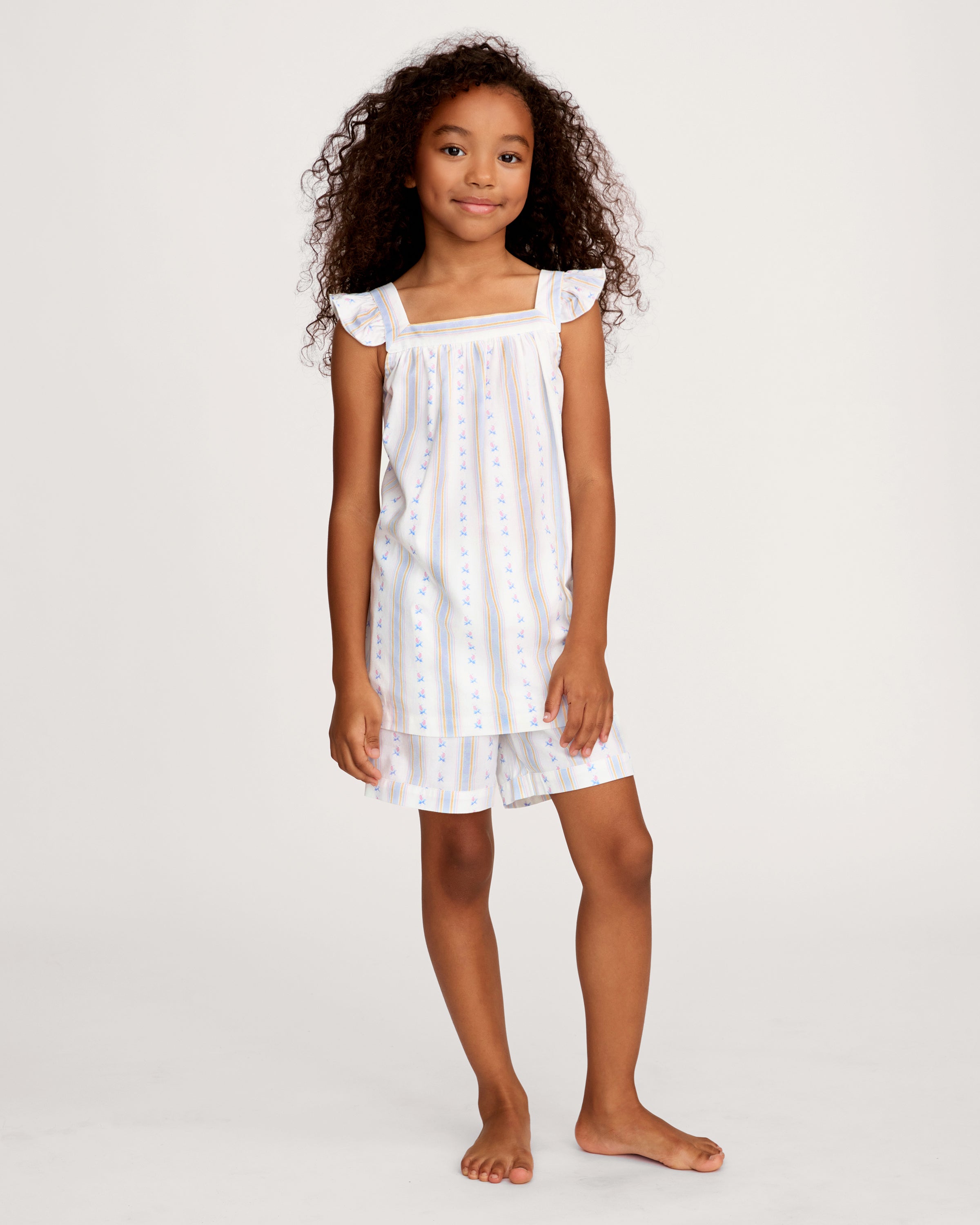 A young girl with curly hair smiles barefoot, wearing the Girl's Twill Clara Short Set in Cottage Stripe— a white sleeveless nightgown and matching shorts—against a plain, light background.