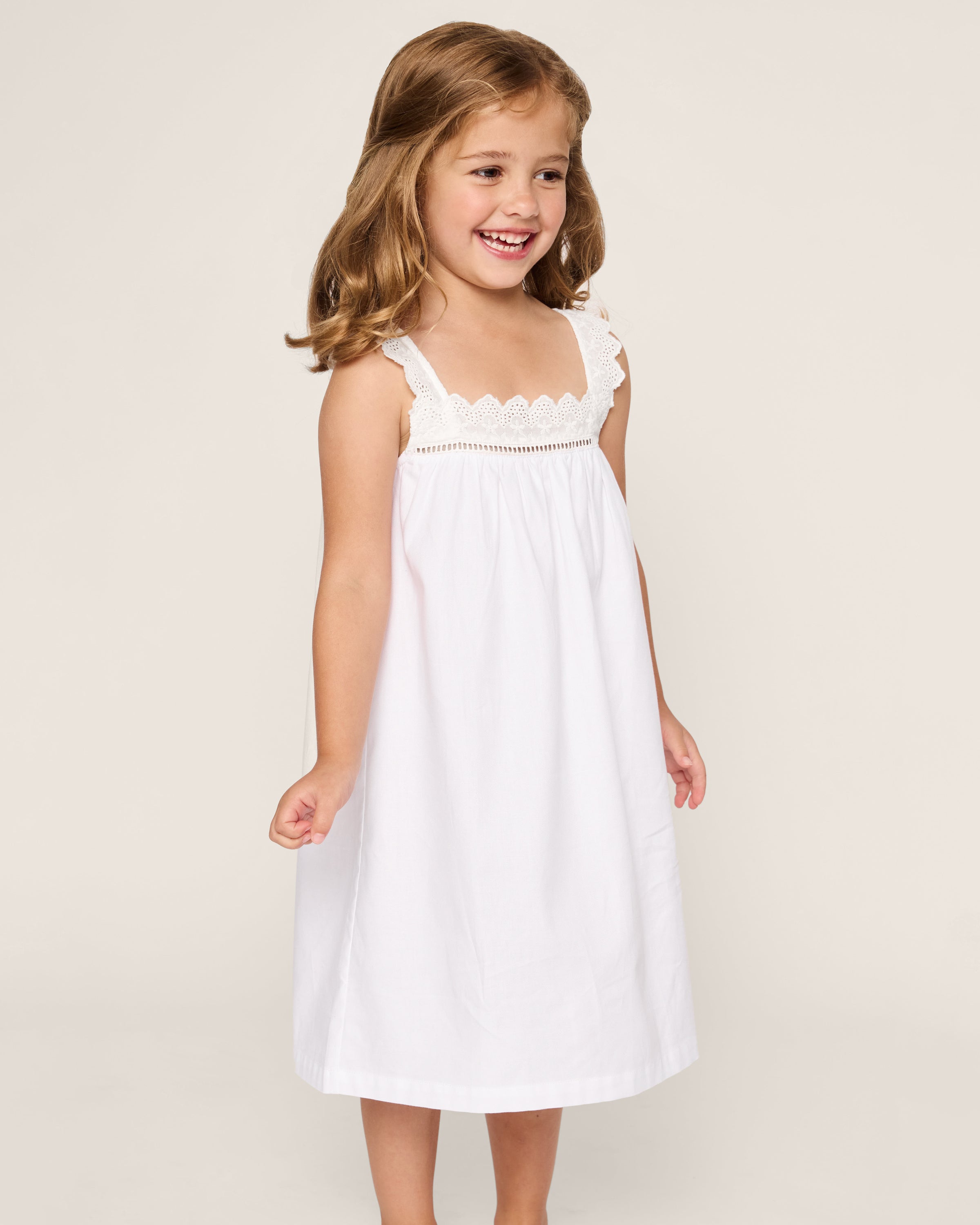 A young girl with brown hair smiles as she looks left, wearing a white sleeveless Girls Twill Esme Nightgown from Petite Plume, featuring lace-detailed straps and made of soft, yarn-dyed cotton. The background is plain and light-colored.