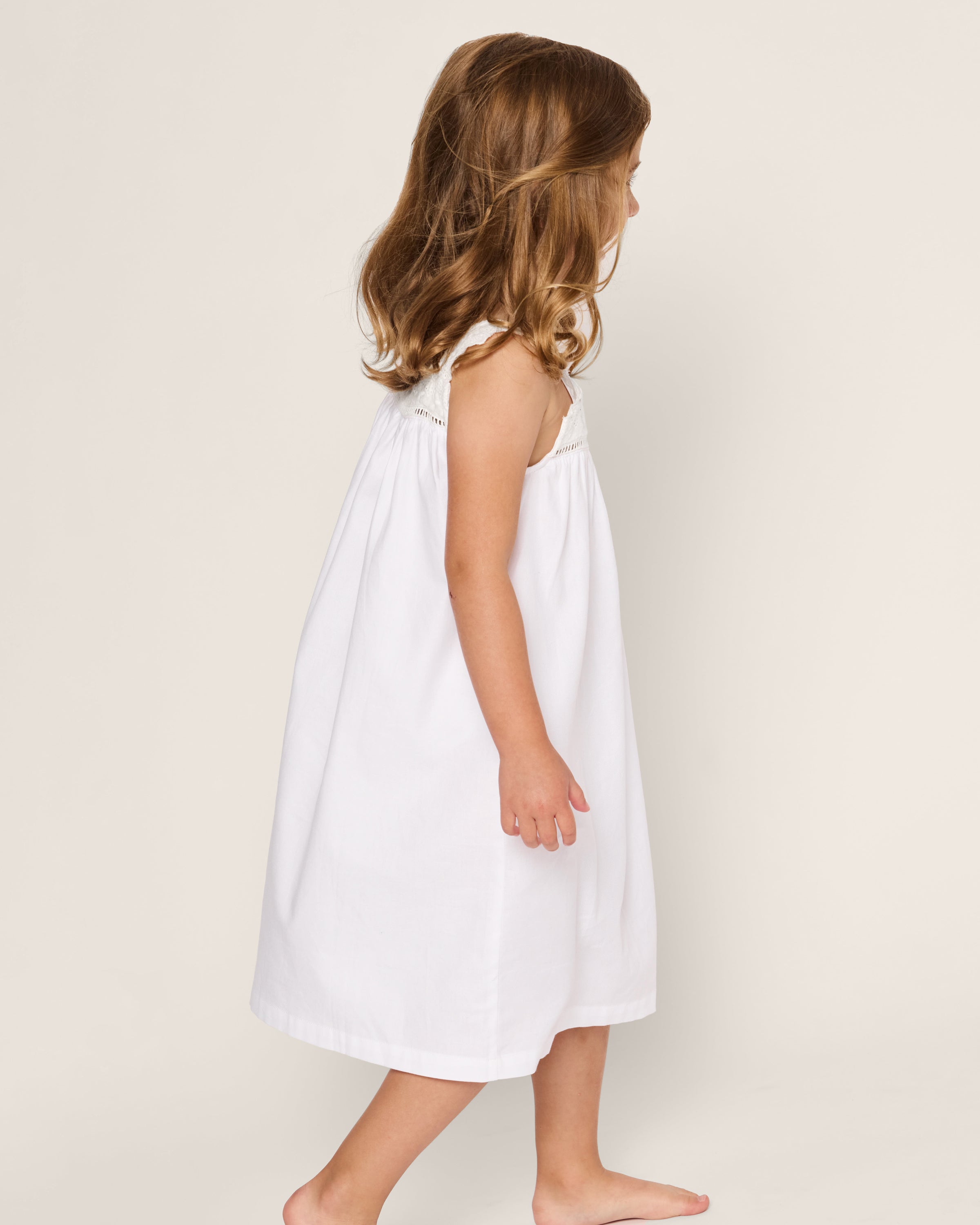A young girl with wavy brown hair is wearing the Petite Plume Girls Twill Esme Nightgown in White. She stands sideways against a plain, light background, barefoot and looking slightly downward.