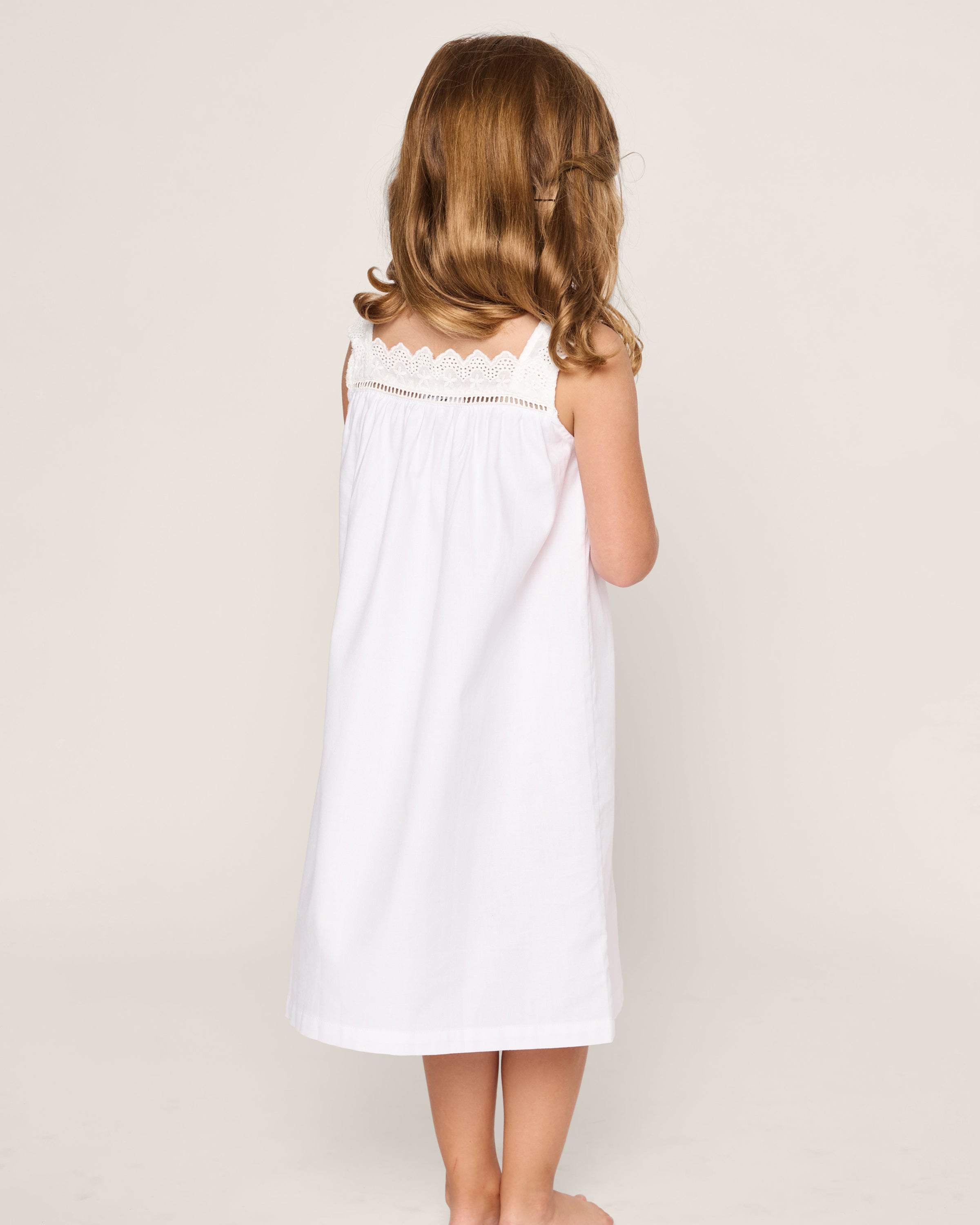A young girl with light brown hair stands barefoot, her back to the camera, wearing Petite Plumes Girls Twill Esme Nightgown in White, featuring lace detail on soft yarn-dyed cotton. The background is a plain, light color.