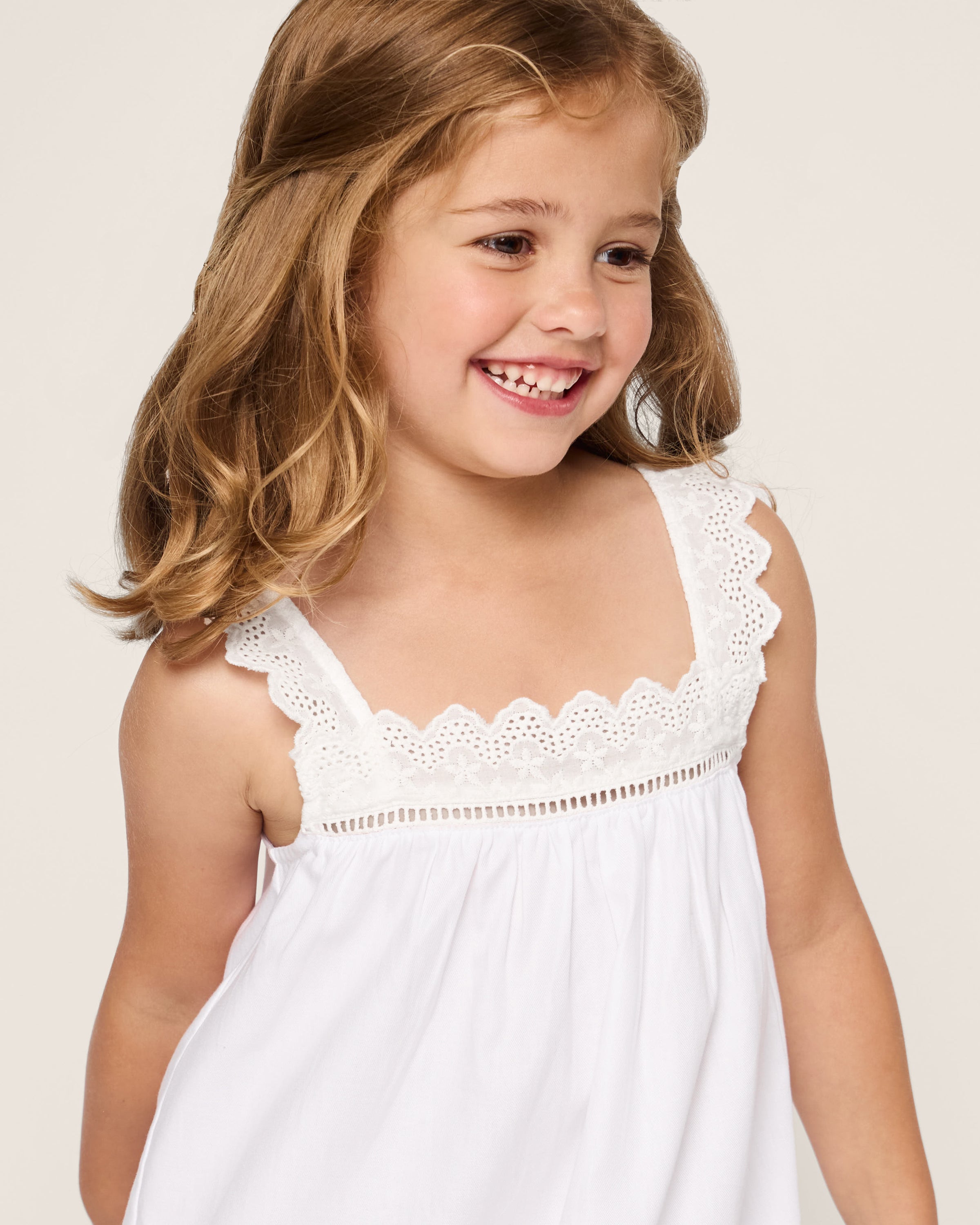 A young girl with shoulder-length blonde hair smiles in Petite Plumes Girls Twill Esme Nightgown in white, featuring lace along the neckline. Made from yarn-dyed cotton, she looks slightly to the side against a plain background.