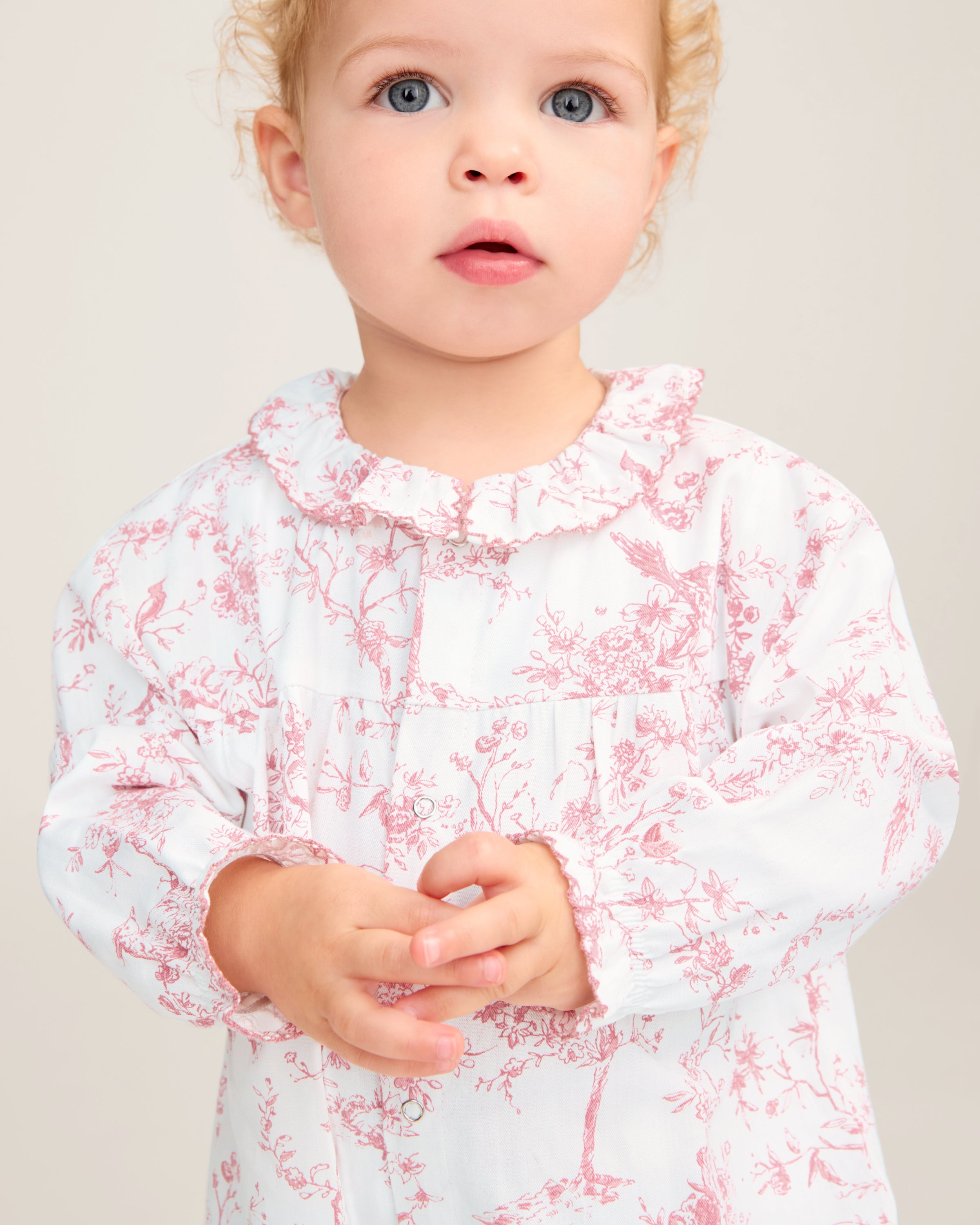 The Baby's Twill Madeline Romper in Pink Timeless Toile features long sleeves, pink toile floral and tree patterns, ruffled cuffs and collar, and convenient inseam snaps for easy diaper changes.