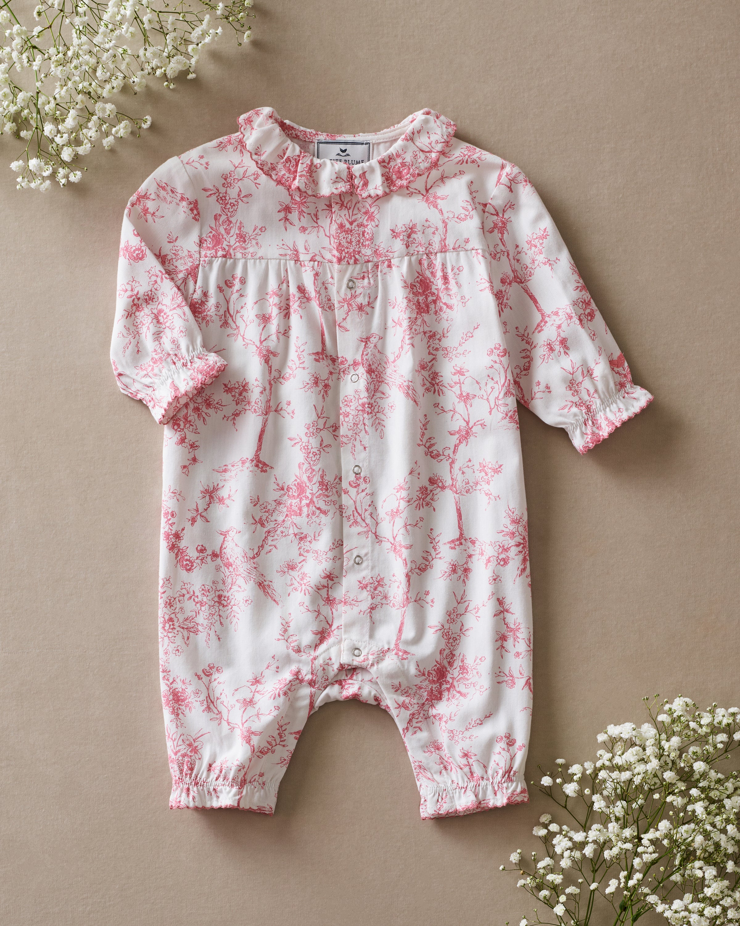 The Baby's Twill Madeline Romper in Pink Timeless Toile features long sleeves, pink toile floral and tree patterns, ruffled cuffs and collar, and convenient inseam snaps for easy diaper changes.