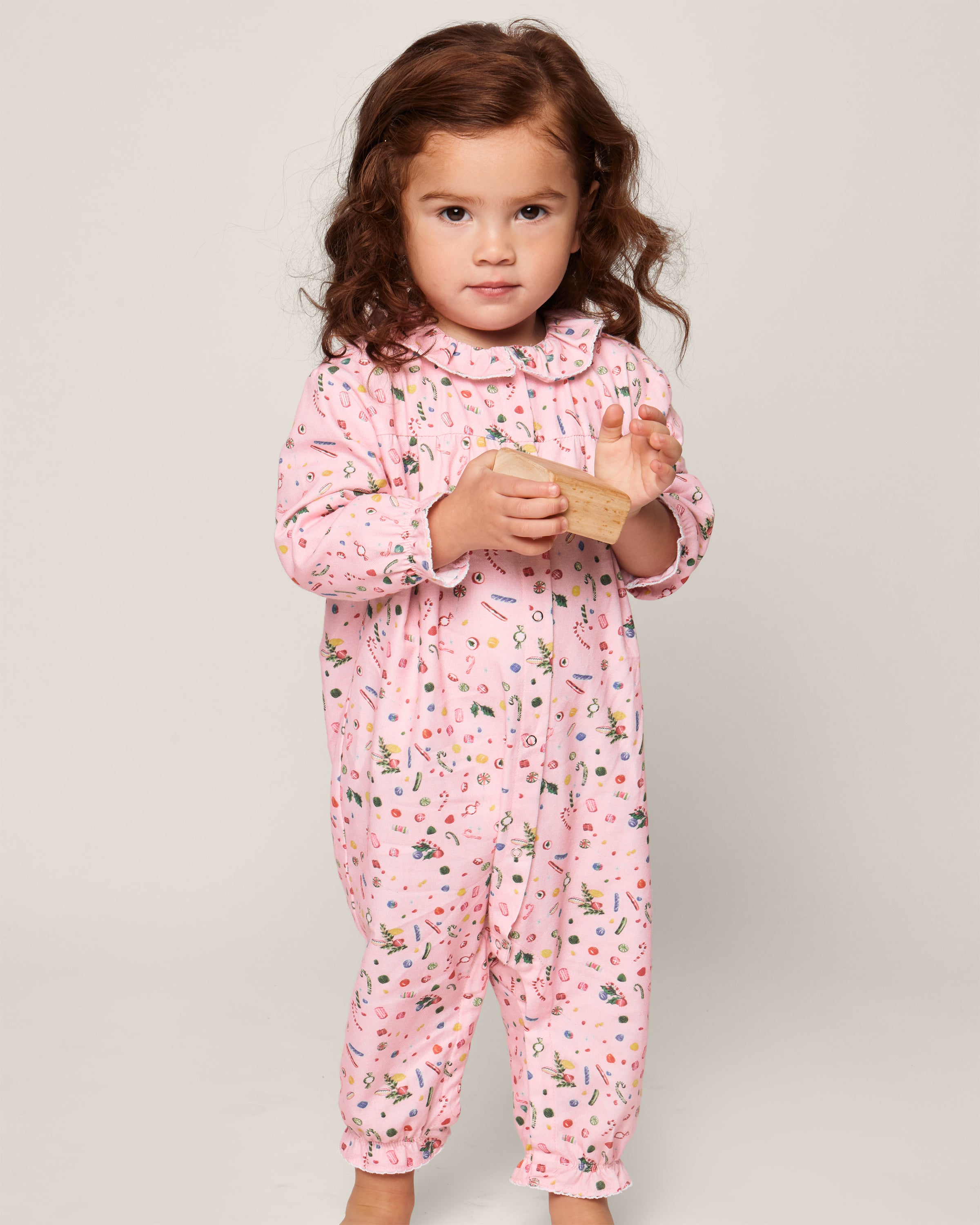 A brown-haired child stands on a plain background wearing Petite Plumes Babys Twill Madeline Romper in Vintage Sweets. The pink onesie with colorful patterns is flame-retardant, and the child holds a wooden object while looking forward.