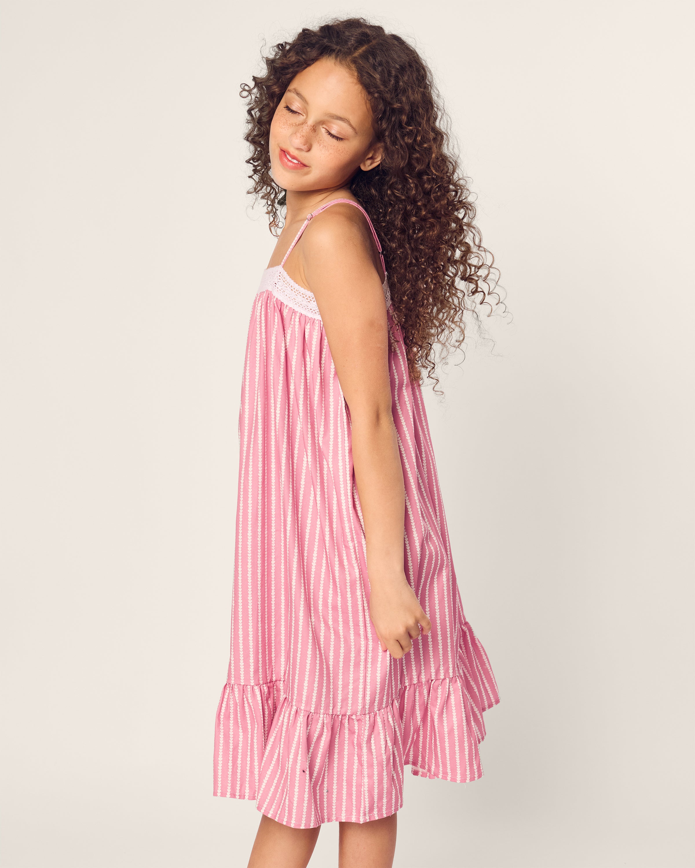The girl with curly hair, wearing a Girls Twill Lilly Nightgown in Love Lines by Petite Plume, stands serenely with eyes closed against a light background.