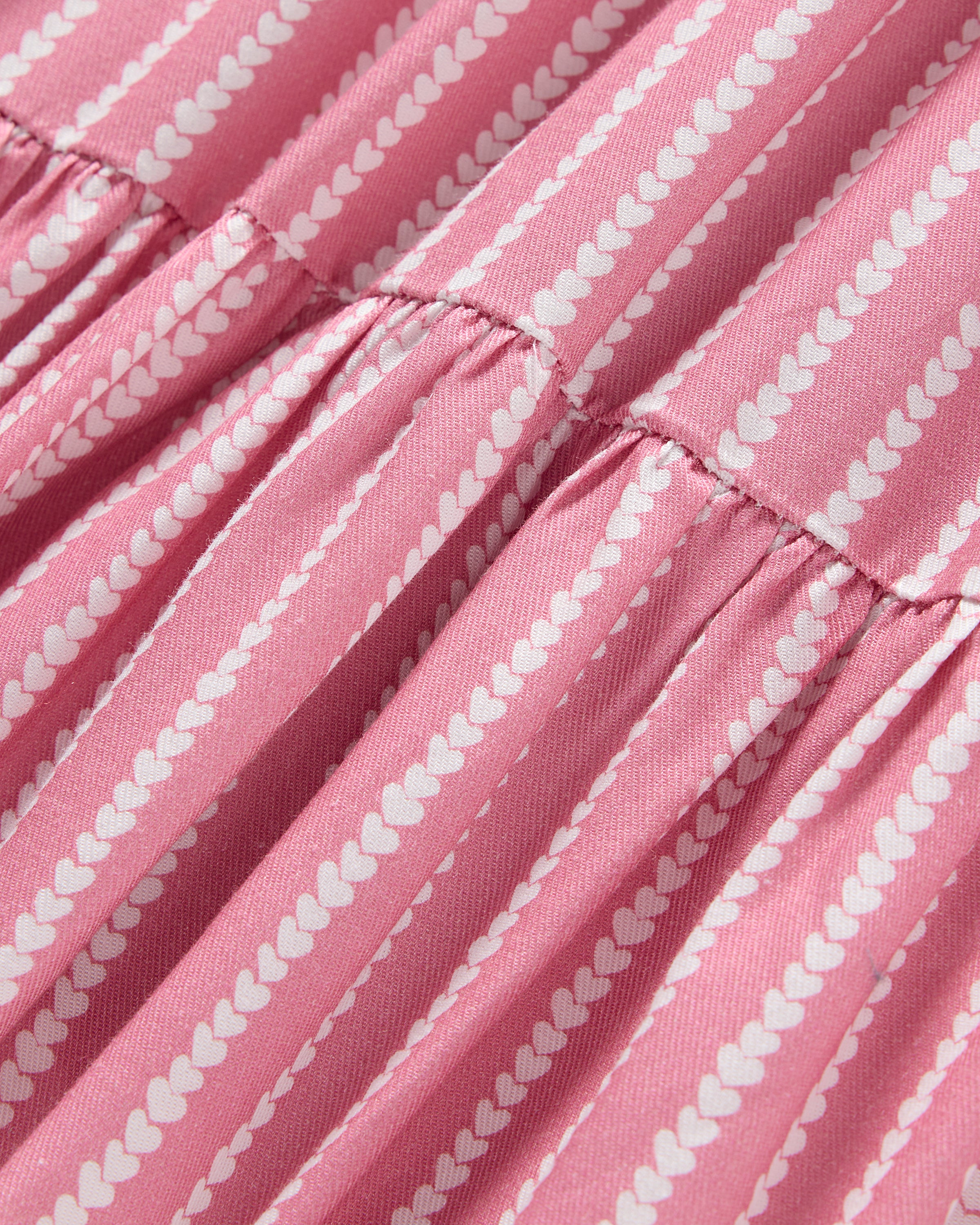 The Girls Twill Lilly Nightgown in Love Lines by Petite Plume features pink and white striped fabric with small heart patterns. The gathered yarn-dyed cotton creates ruffled folds, perfect for luxurious sleepwear.