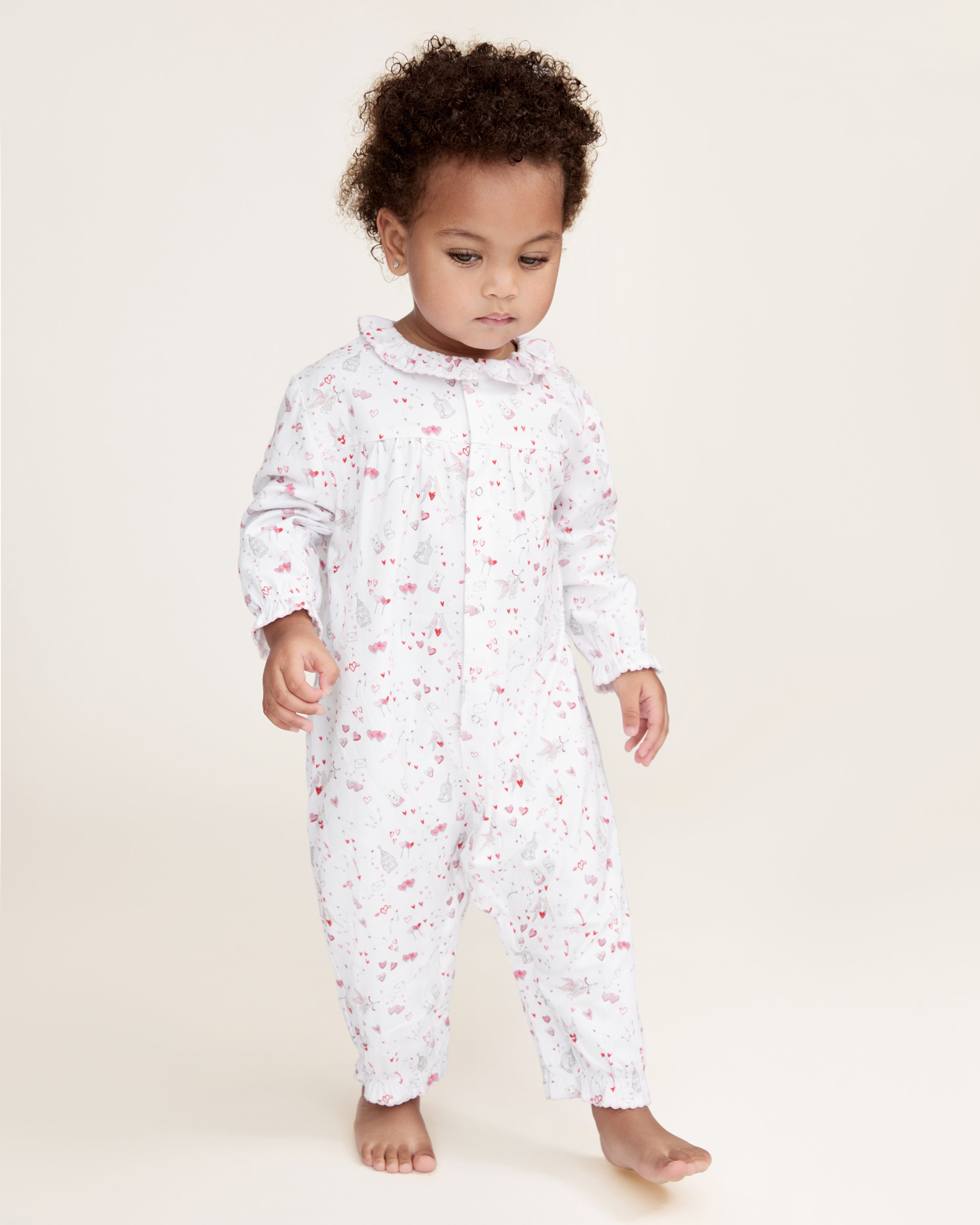 The Baby's Twill Madeline Romper in Love Song is a white long-sleeve romper with ruffled cuffs and collar, decorated with a pink pattern of hearts, glasses, and small animals. It features snap buttons down the front and legs.