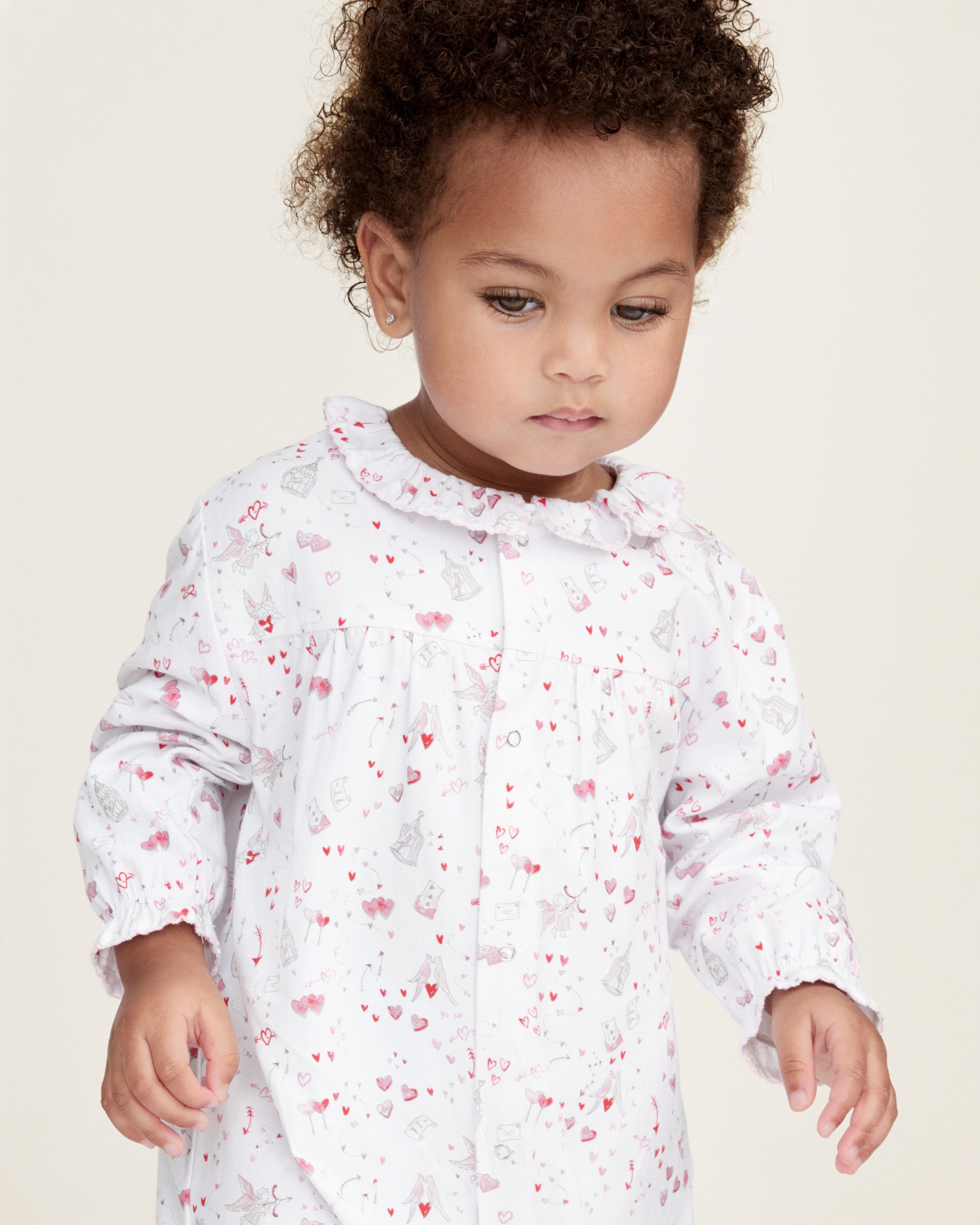 The Baby's Twill Madeline Romper in Love Song is a white long-sleeve romper with ruffled cuffs and collar, decorated with a pink pattern of hearts, glasses, and small animals. It features snap buttons down the front and legs.