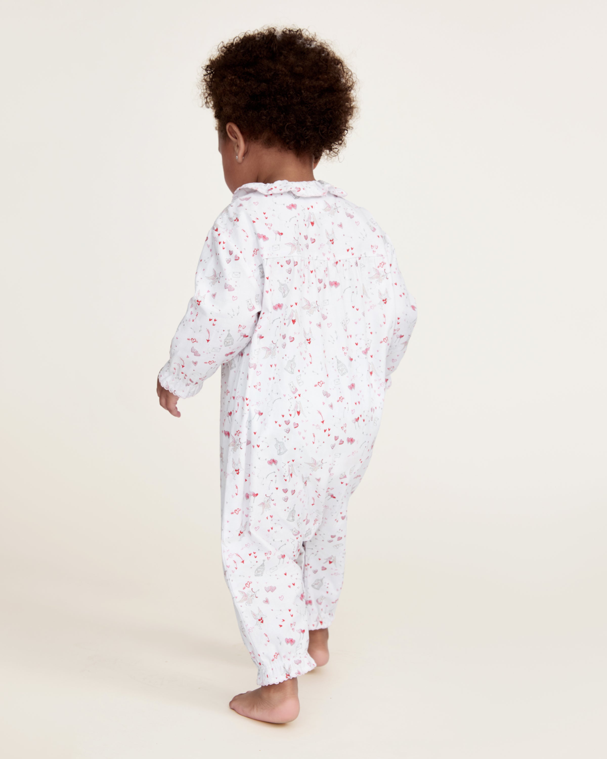 The Baby's Twill Madeline Romper in Love Song is a white long-sleeve romper with ruffled cuffs and collar, decorated with a pink pattern of hearts, glasses, and small animals. It features snap buttons down the front and legs.