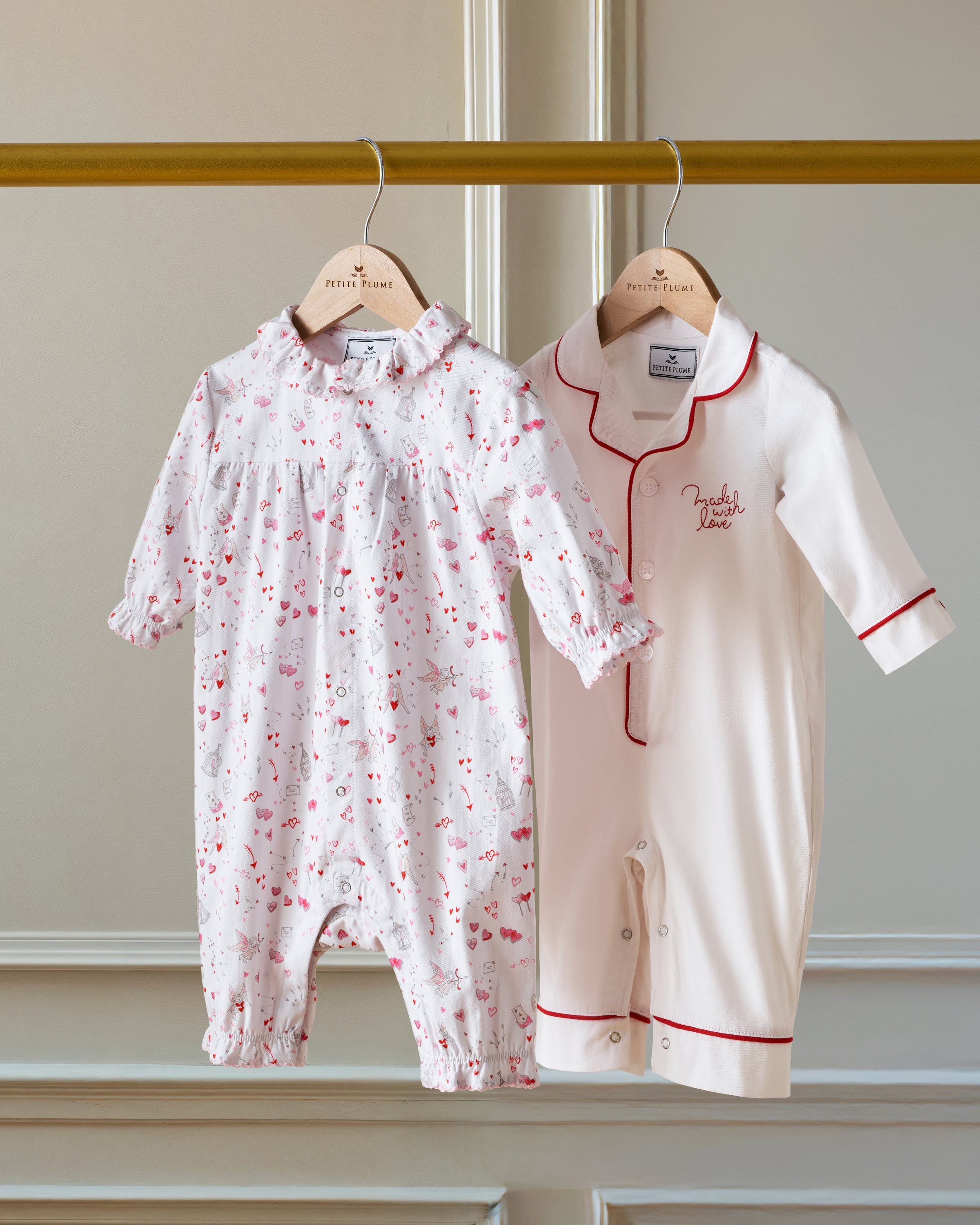 The Baby's Twill Madeline Romper in Love Song is a white long-sleeve romper with ruffled cuffs and collar, decorated with a pink pattern of hearts, glasses, and small animals. It features snap buttons down the front and legs.