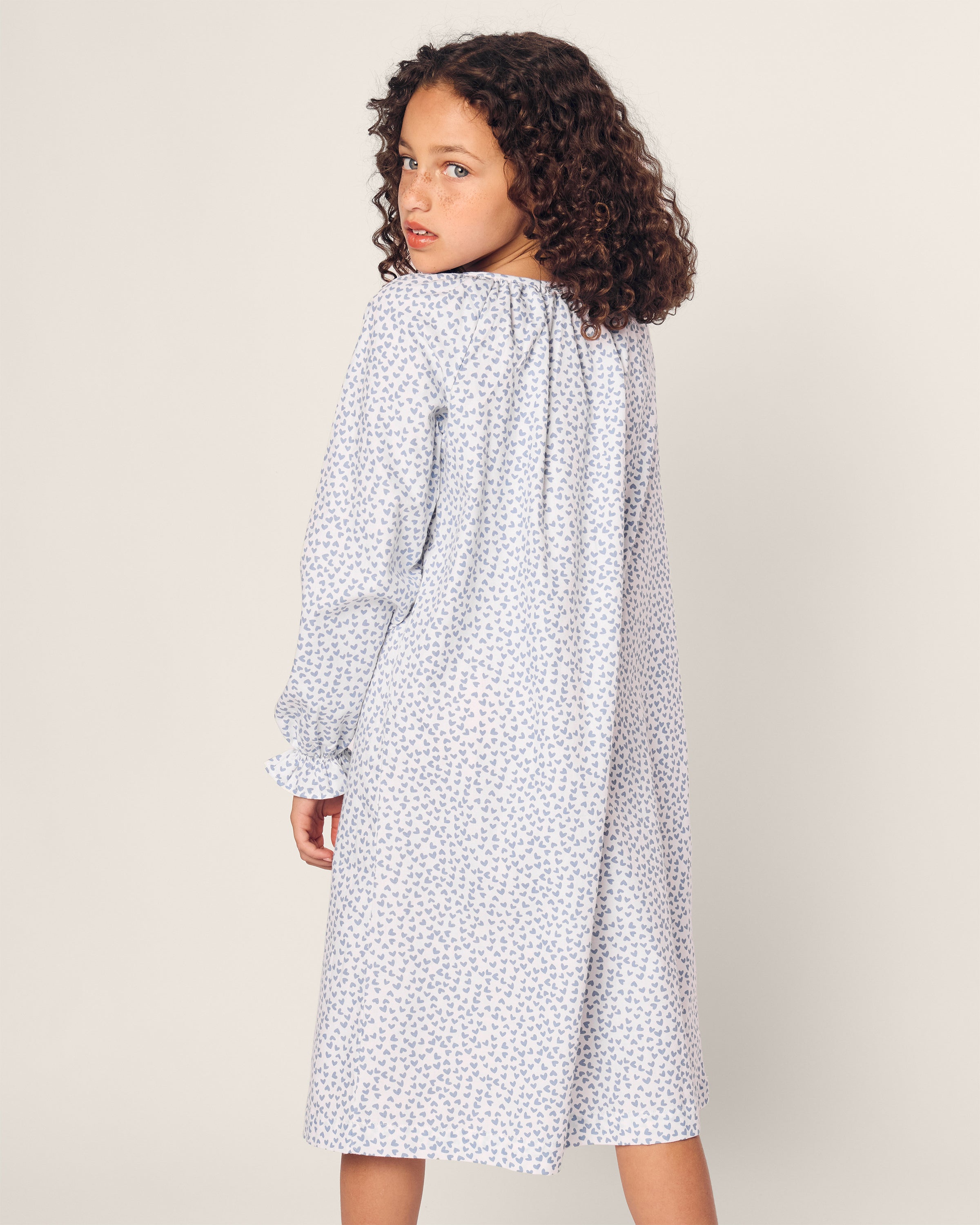 A young girl with curly hair wears Petite Plumes Girls Twill Delphine Nightgown in Bluehearts, looking over her shoulder against a plain background.