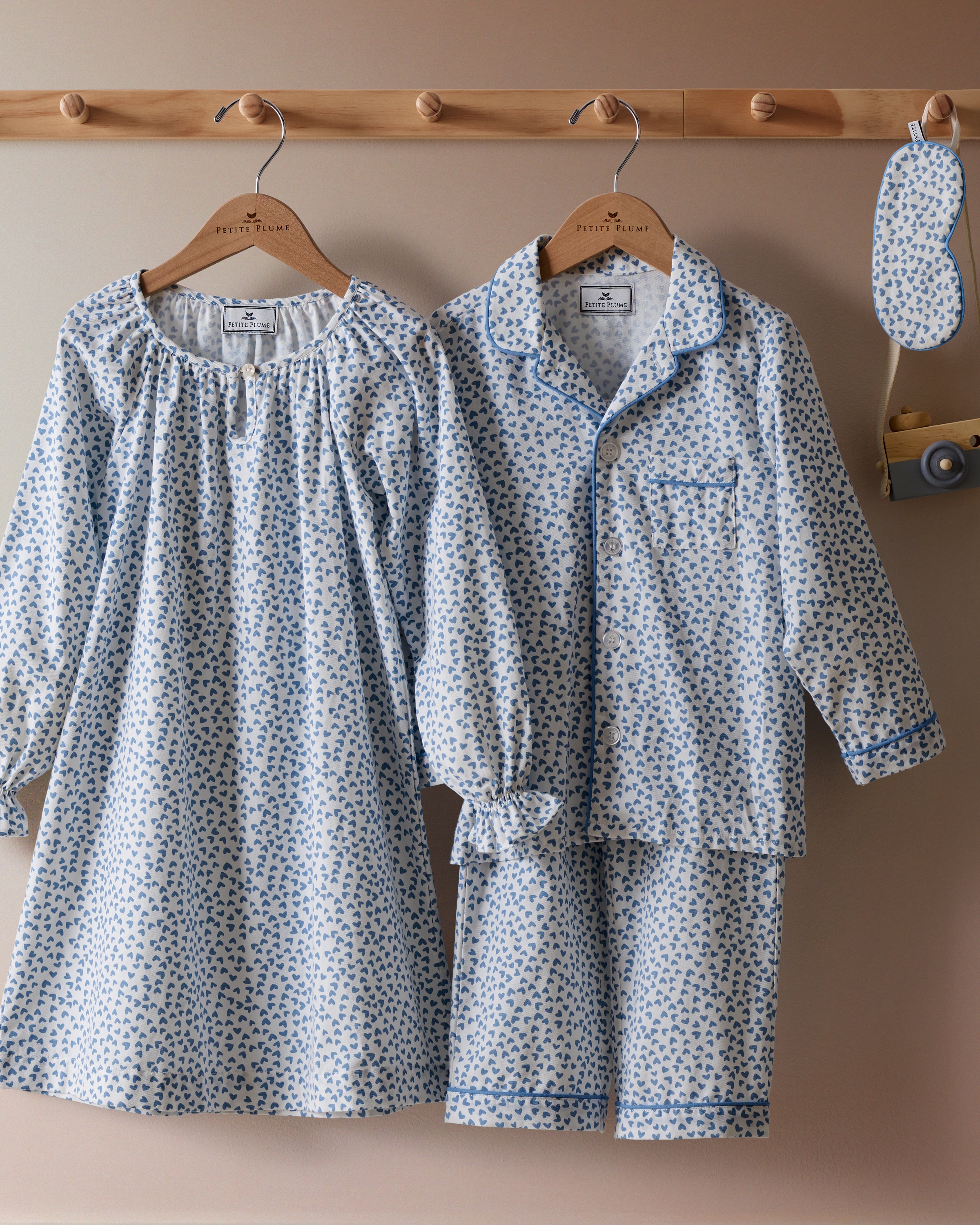 Two Petite Plume luxurious sleepwear sets with a Bluehearts design hang on a wooden rod: the elegant Girls Twill Delphine Nightgown, and a chic button-up shirt with shorts. An eye mask complements the ensemble, inviting restful slumber.