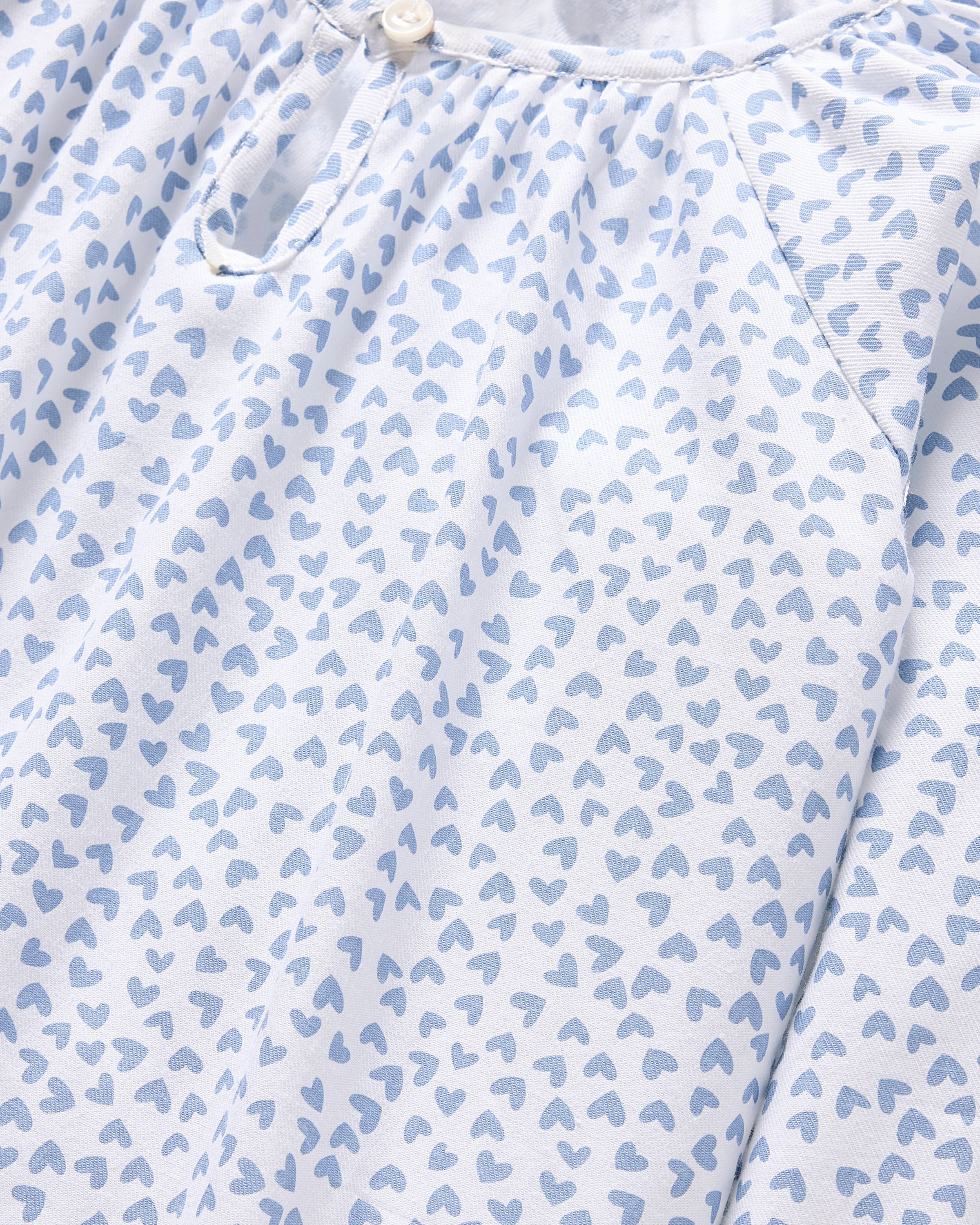 Close-up of a white fabric with a pattern of small, light blue hearts. The Petite Plume Girls Twill Delphine Nightgown in Bluehearts features a partially visible buttoned placket at the top, suggesting its part of luxurious sleepwear adorned with the charming Bluehearts design.