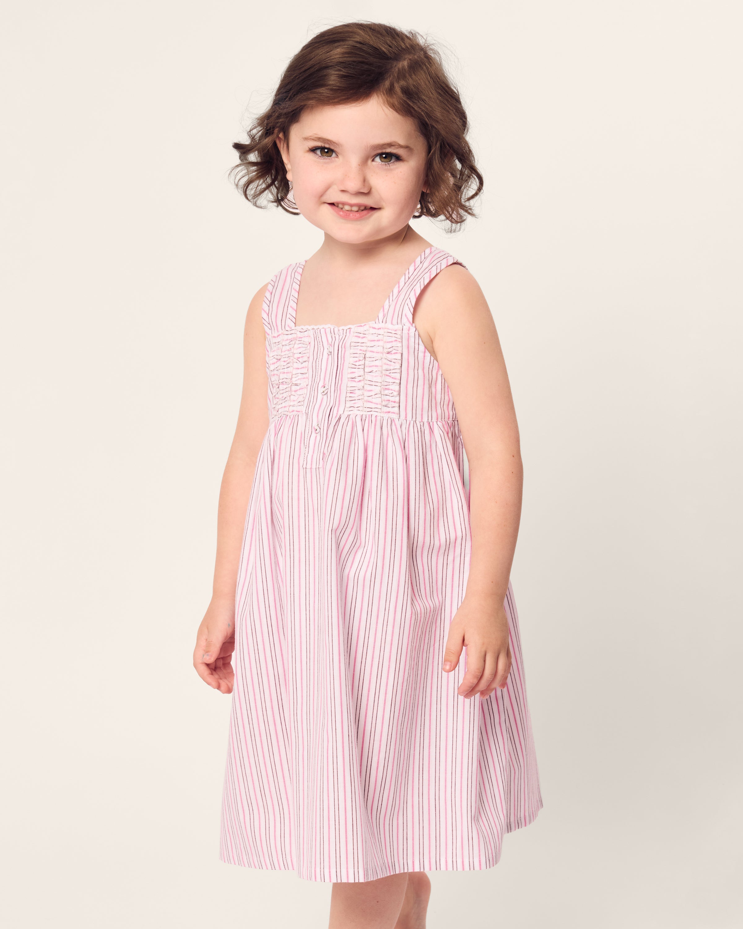 A young girl with short brown hair wears the charming Petite Plume Girls Twill Charlotte Nightgown in Blush Boulevard. She smiles while standing against a simple, light-colored background.