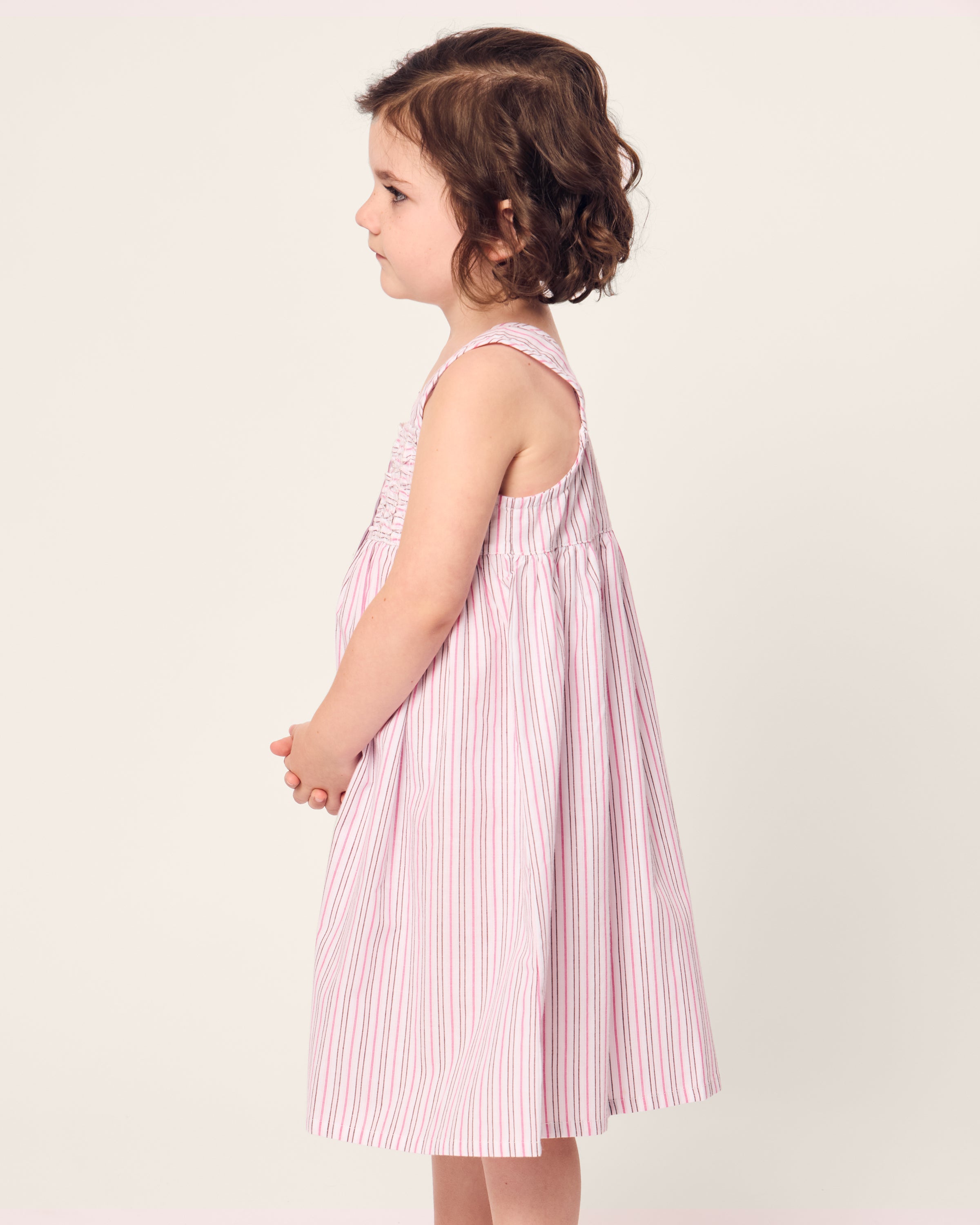 A young child with short, curly brown hair stands in profile, facing left, wearing a Petite Plume Girls Twill Charlotte Nightgown in Blush Boulevard. The background is a plain off-white.