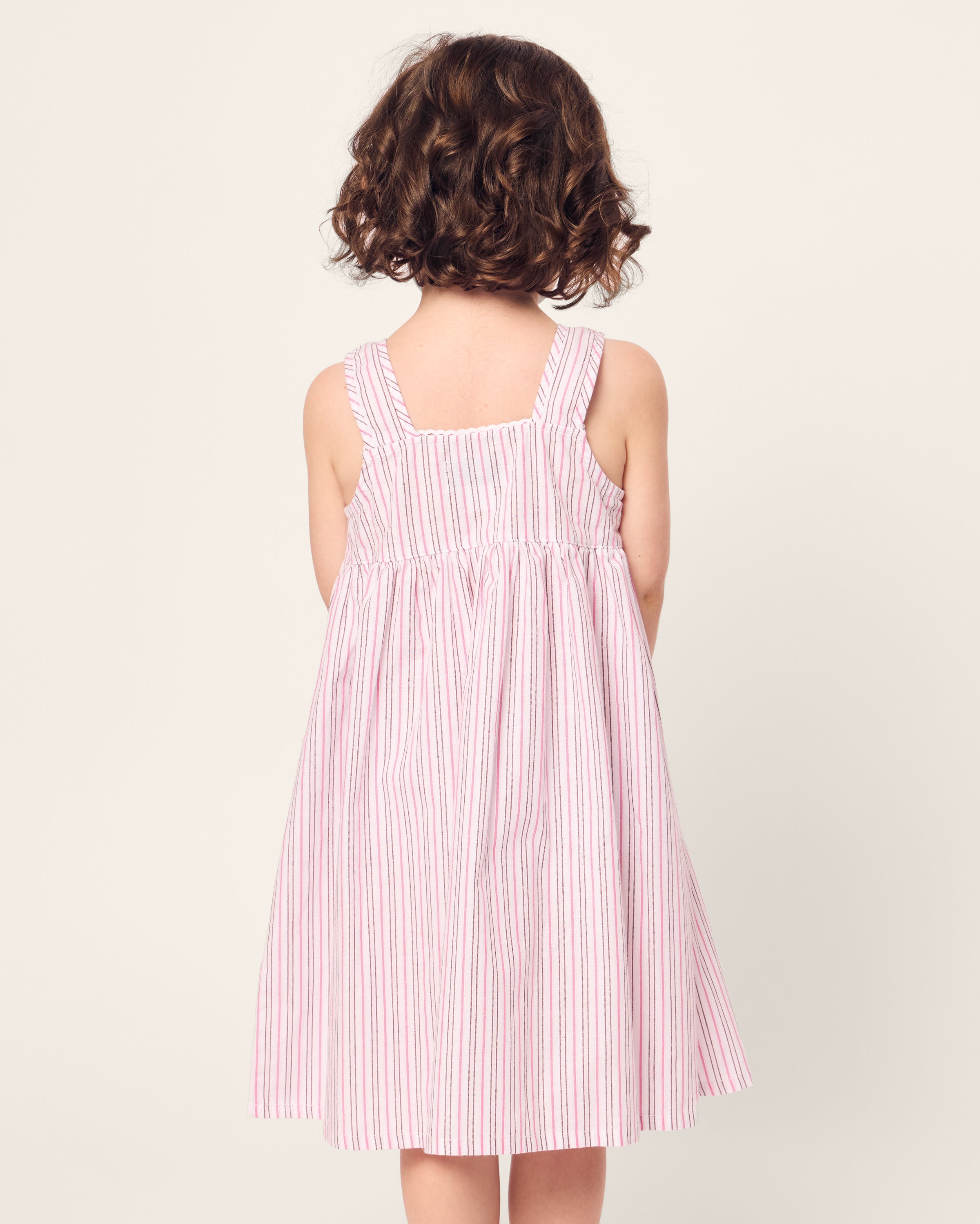A young child with curly brown hair stands with their back to the camera, wearing a Petite Plume Girls Twill Charlotte Nightgown in Blush Boulevard. The background is plain and light-colored.