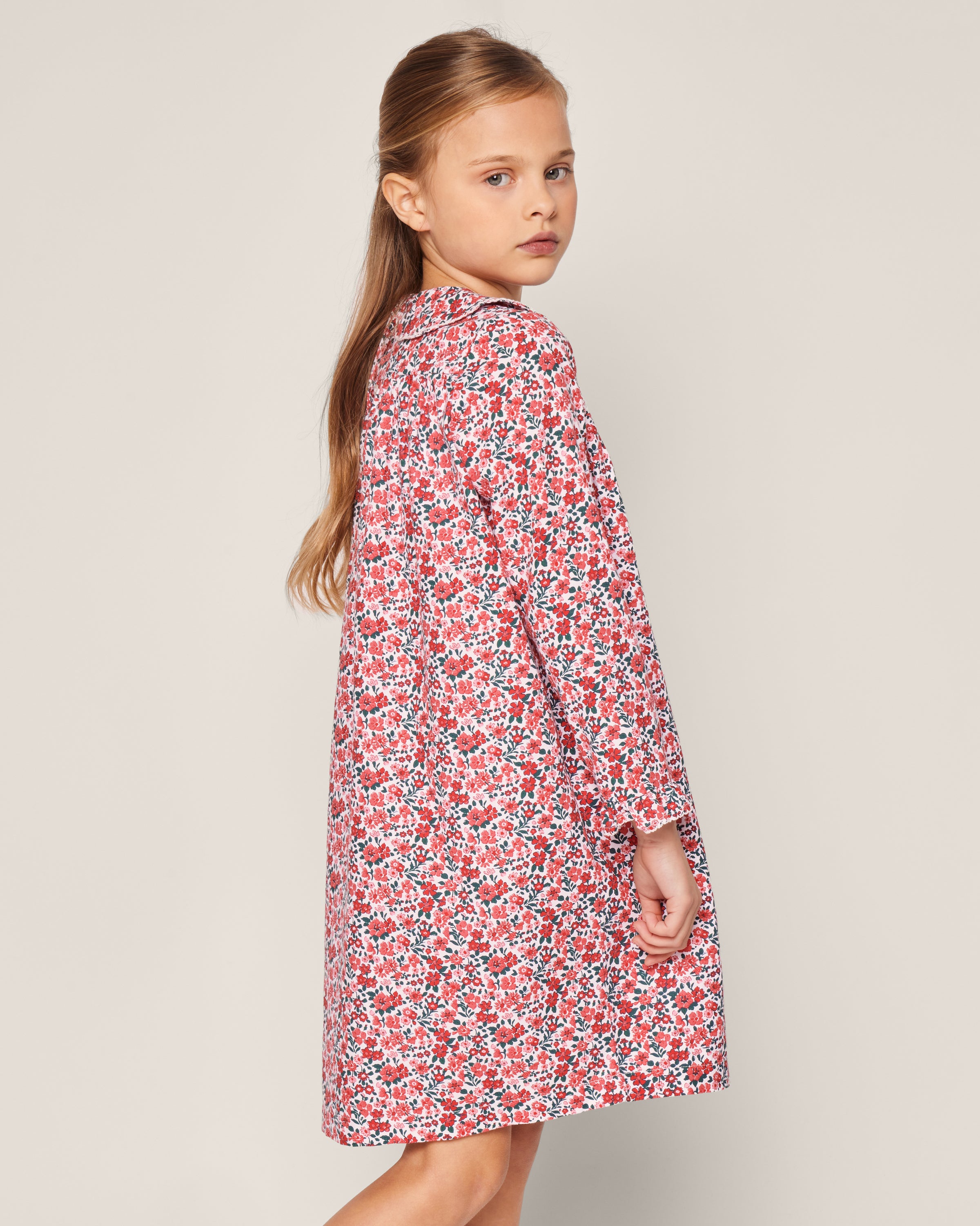 A young girl with long brown hair stands sideways, looking over her shoulder. Shes wearing Petite Plumes Girls Eloise Nightgown in Fleurs des Bois, a floral print nightgown adorned with red and white blooms on a light background, exuding graceful charm.