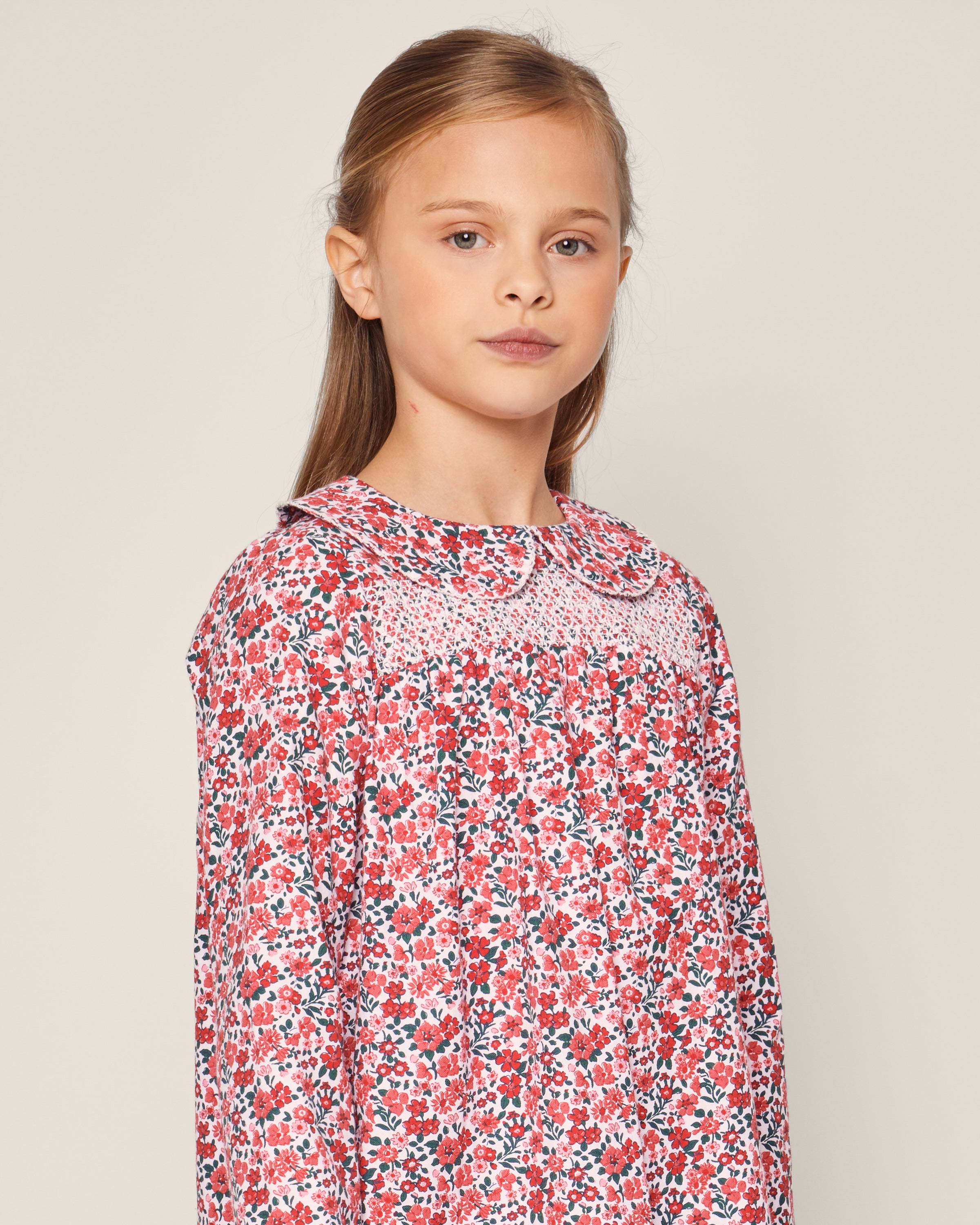 A young girl with long brown hair wears Petite Plumes Girls Eloise Nightgown in Fleurs des Bois, covered with red and pink flowers. She stands against a plain, light background, gazing slightly to the side. The nightgown is designed with a Peter Pan collar and gathered detailing.