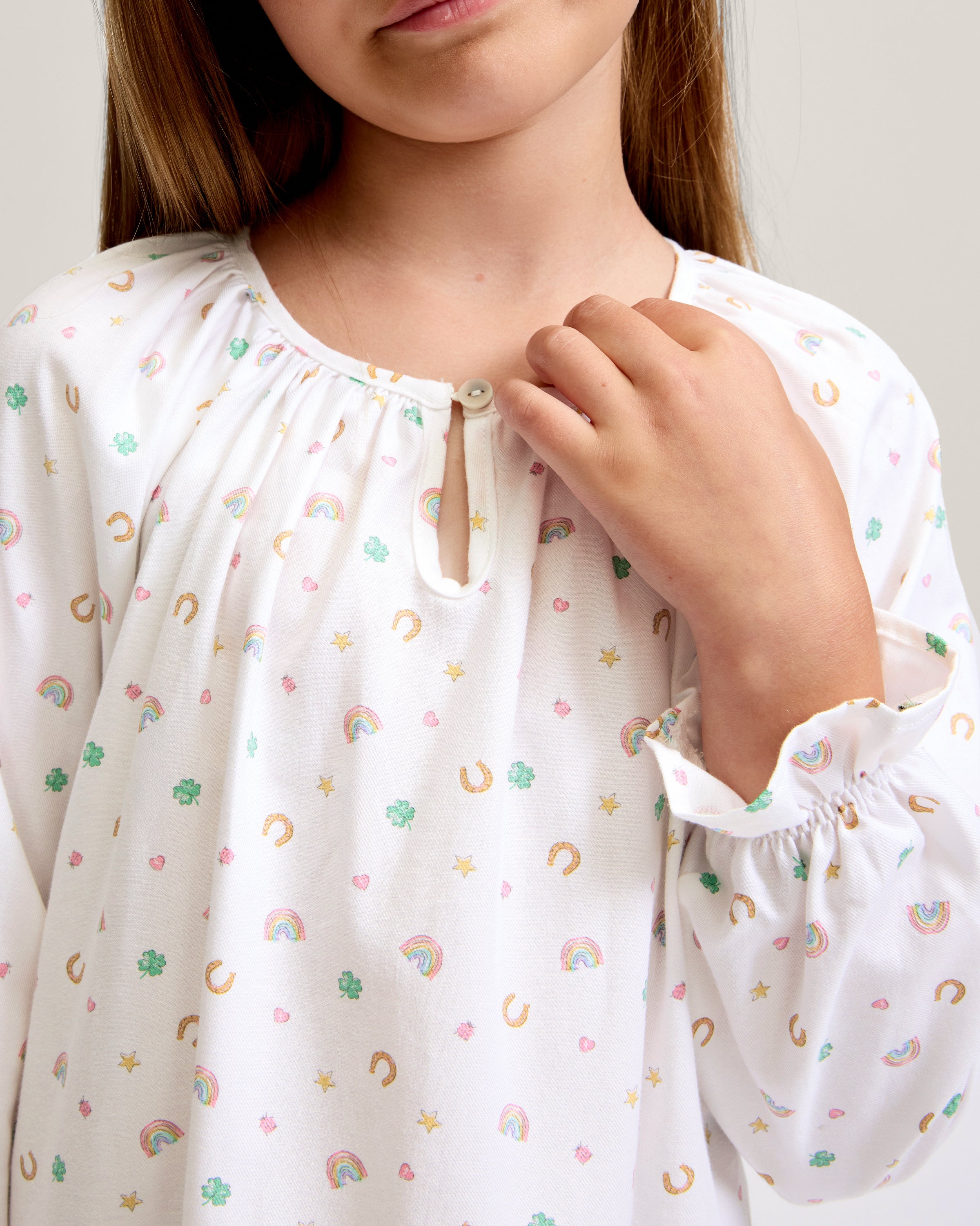 A young girl stands barefoot on a light background, wearing the Girl's Twill Delphine Nightgown in Good Luck Charms. She gazes upward thoughtfully, showcasing its long sleeves and colorful pattern.