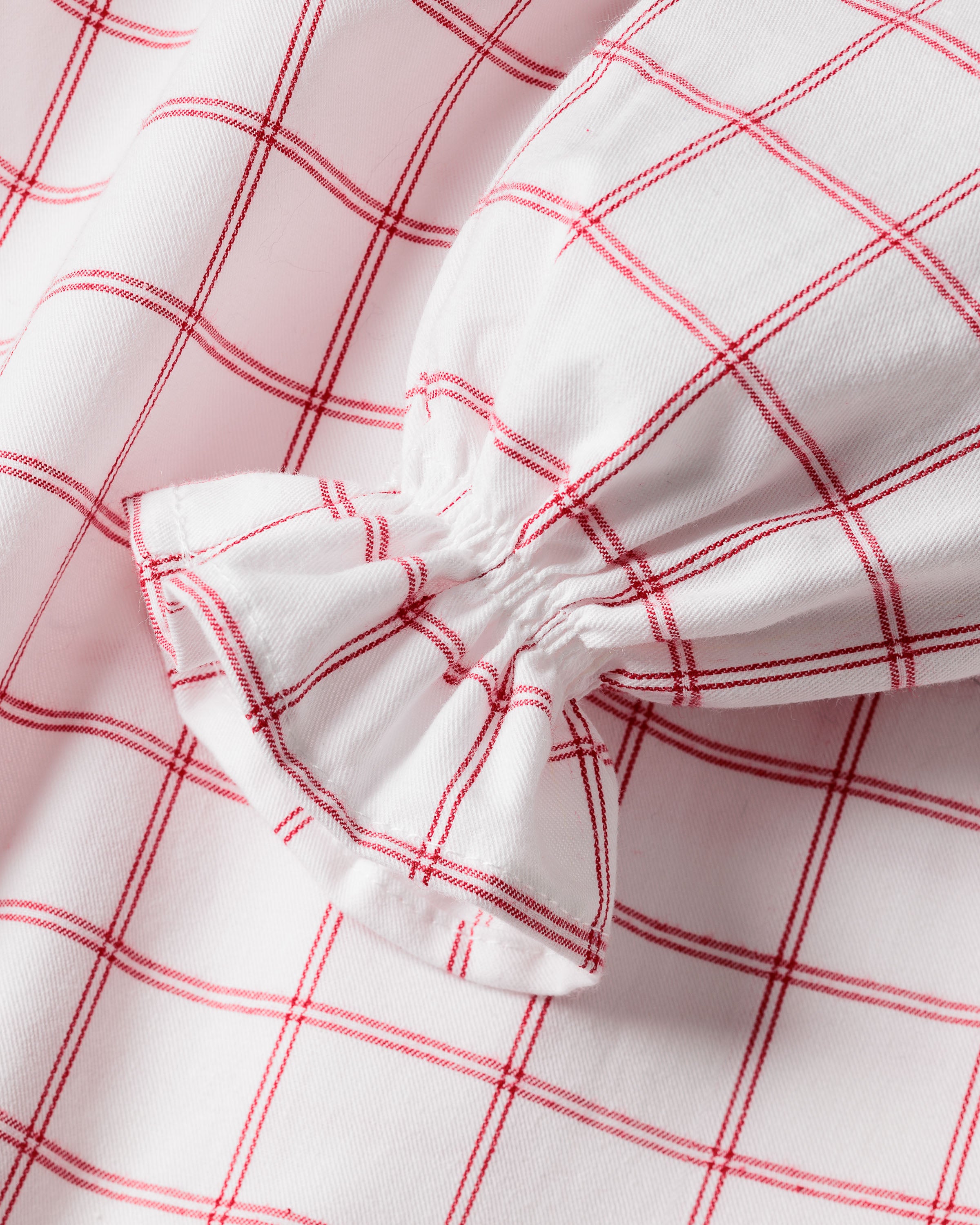 Close-up of a fabric featuring the garnet-red and white checkered pattern of Petite Plumes Girls Twill Delphine Nightgown in Garnet Tattersall. The material is gathered or elasticized, creating a ruffled effect with intersecting lines at right angles.