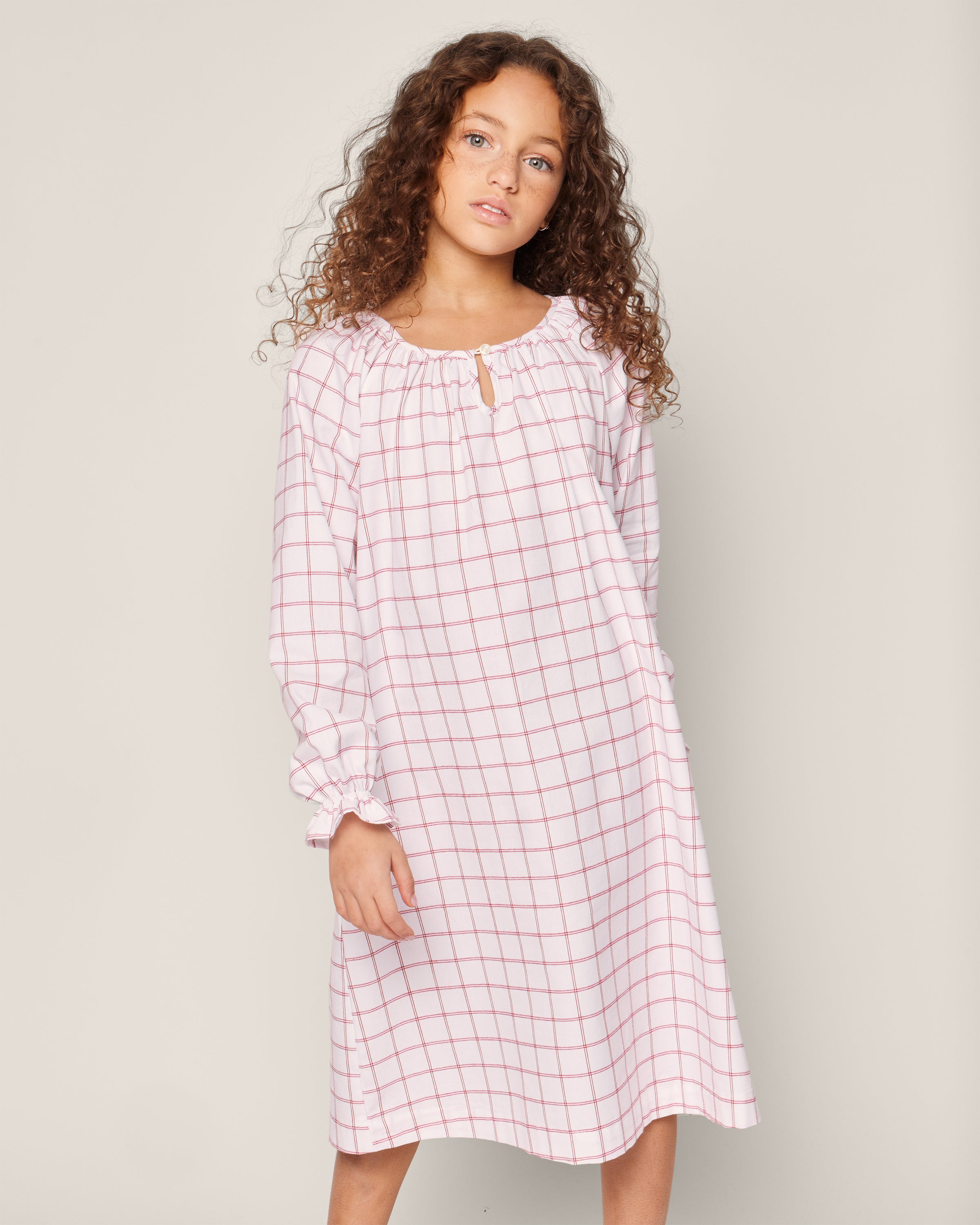 A child with long curly hair wears Petite Plumes Girls Twill Delphine Nightgown in Garnet Tattersall, made from flame-retardant fabric. This white nightgown features a pink checkered pattern, long sleeves, and a gathered neckline against a plain light-colored background.