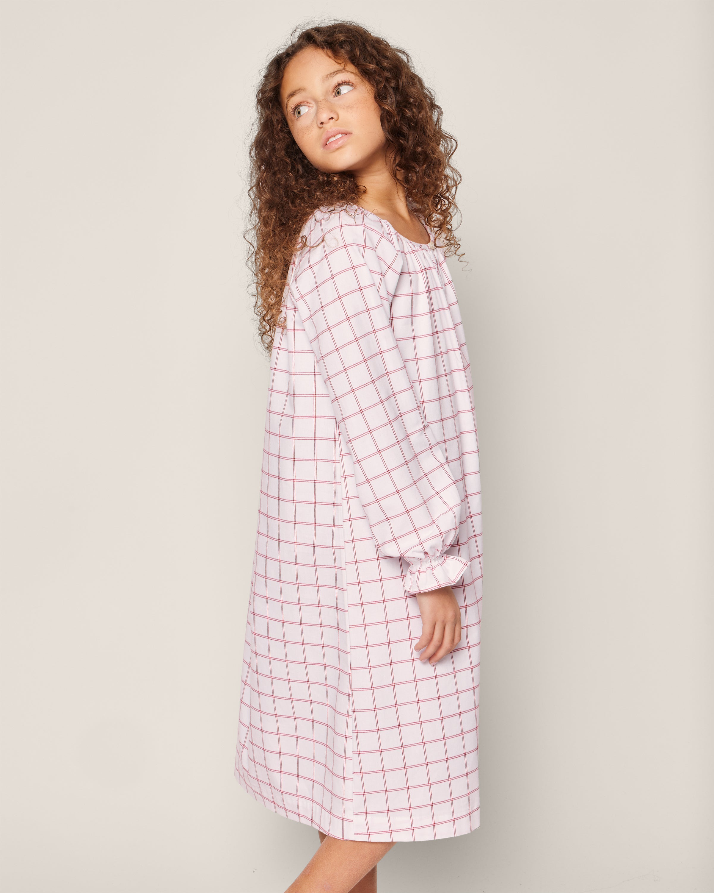 A young person with curly hair wears the Petite Plume Girls Twill Delphine Nightgown in Garnet Tattersall. They stand in profile against a plain background, looking over their shoulder.