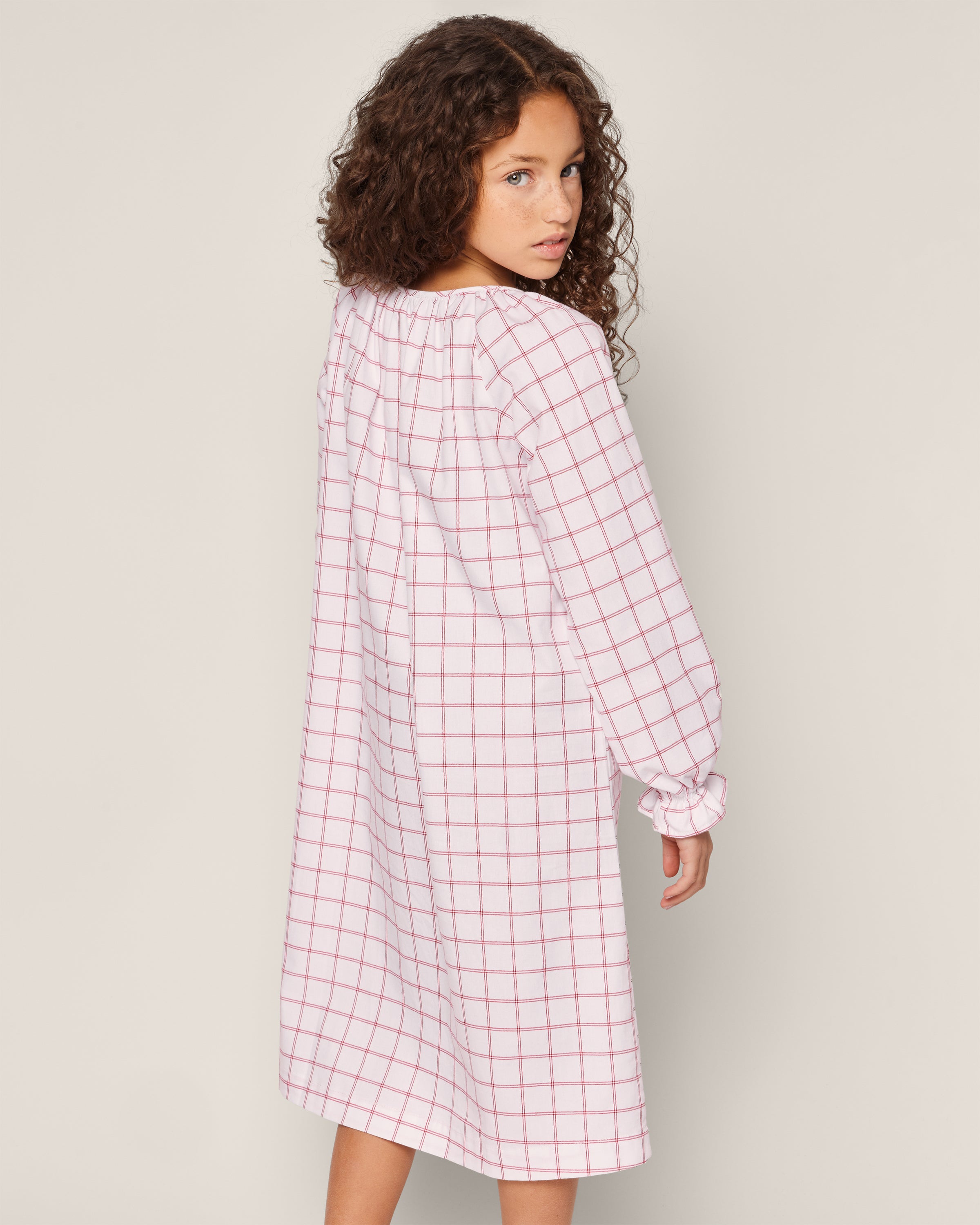 A child in Petite Plumes Girls Twill Delphine Nightgown in Garnet Tattersall, featuring long sleeves and flame-retardant fabric, stands against a plain background, glancing over their shoulder.