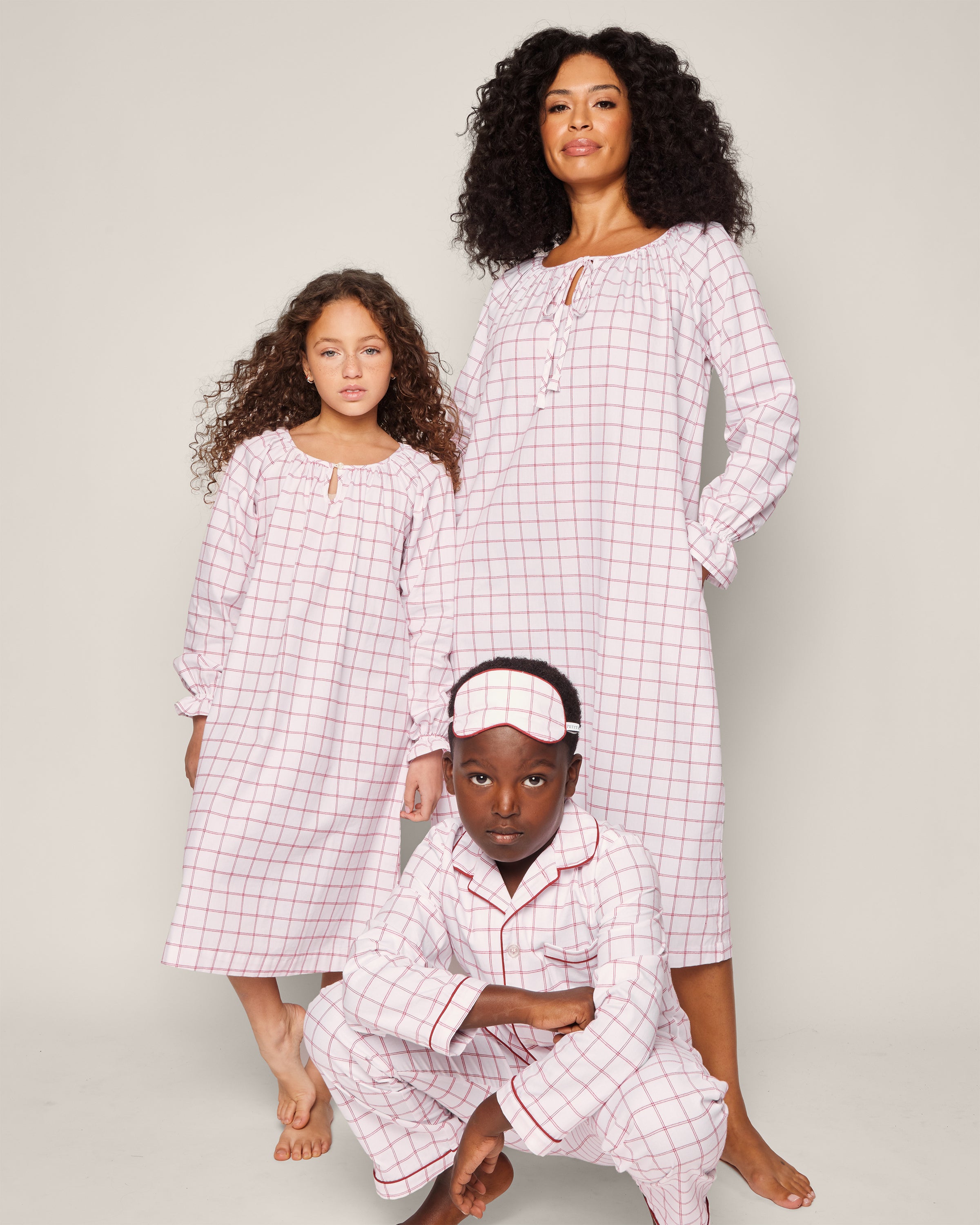 Three people in matching garnet-red checkered pajamas pose together. An adult stands as a child peeks from behind; another crouches with a matching sleep mask. This scene spotlights Petite Plumes Girls Twill Delphine Nightgown in Garnet Tattersall, crafted from flame-retardant fabric against a plain background.