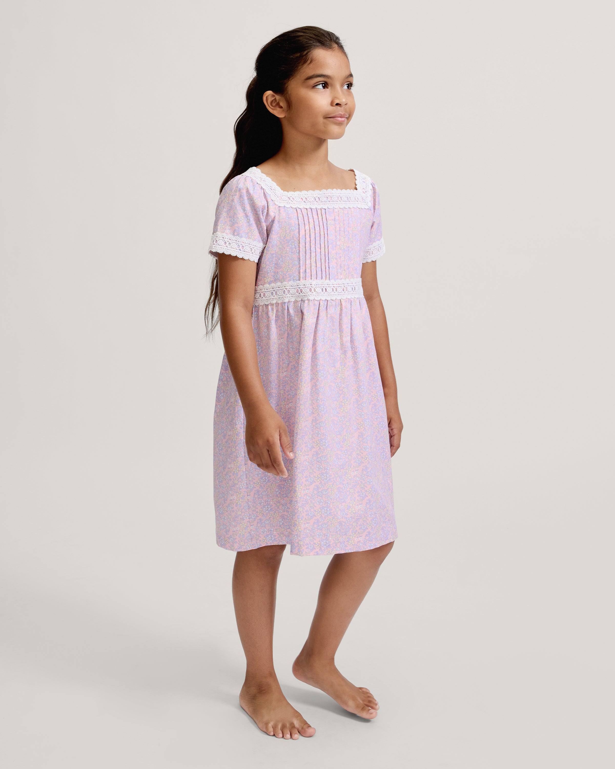 A young girl with long dark hair stands barefoot, wearing the Girl's Twill Juliette Nightgown in Fleur Blush—a light purple, knee-length dress featuring short sleeves, lace trim, and a delicate floral pattern—against a plain light background.