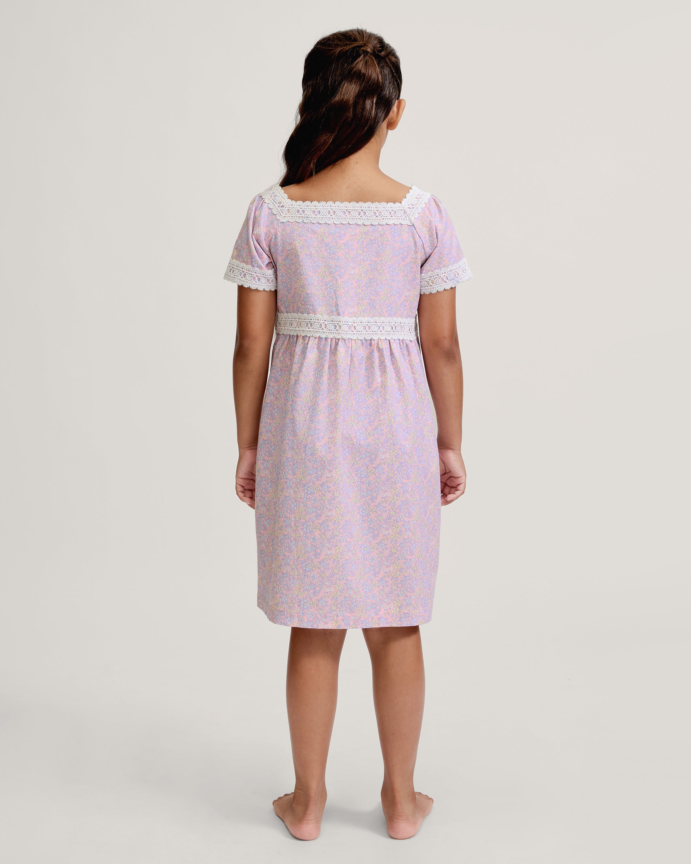 A young girl with long dark hair stands barefoot, wearing the Girl's Twill Juliette Nightgown in Fleur Blush—a light purple, knee-length dress featuring short sleeves, lace trim, and a delicate floral pattern—against a plain light background.