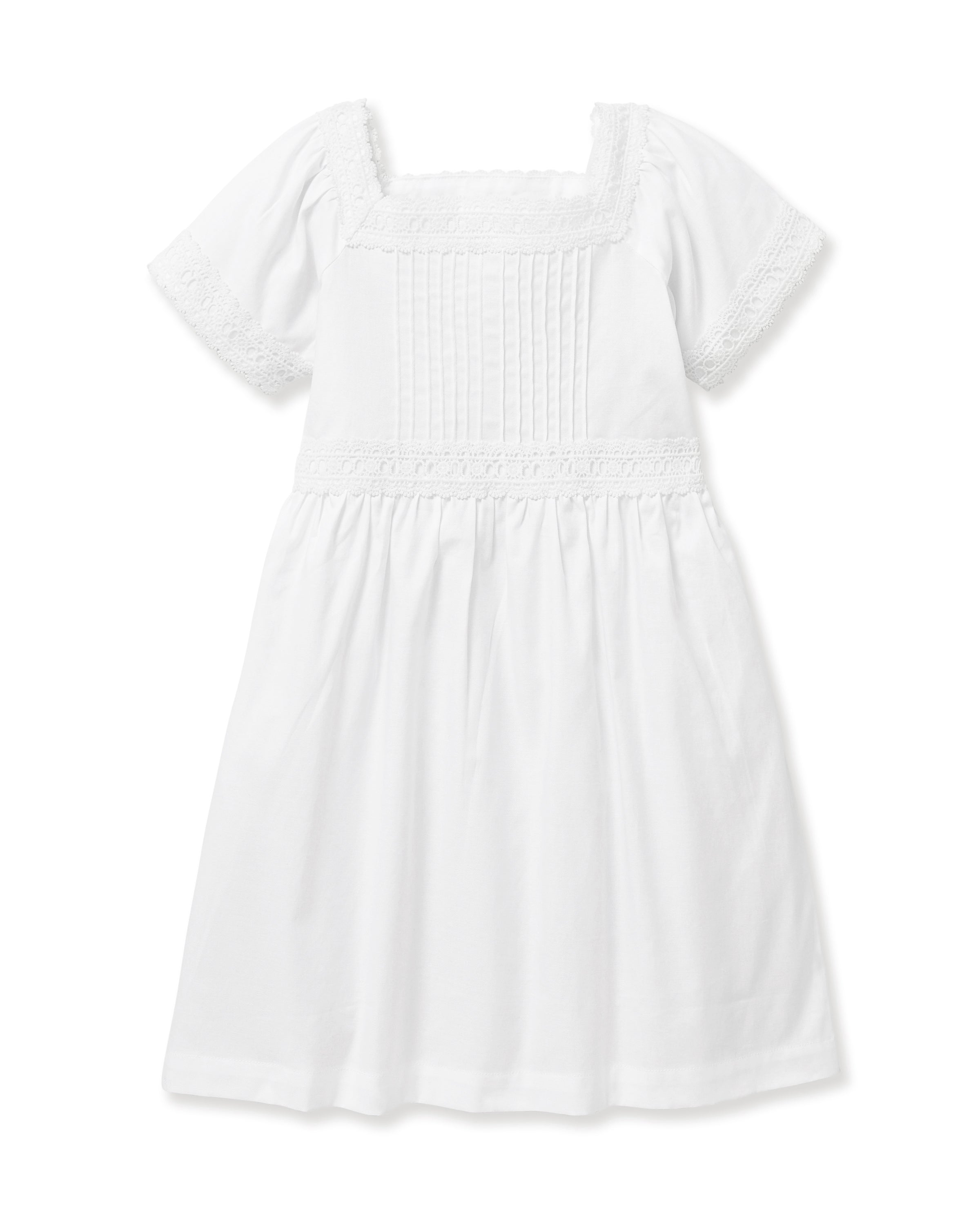 A young girl stands barefoot against a light background, wearing the Girl's Twill Juliette Nightgown in White—a knee-length piece with short sleeves, lace trim, and pintuck details. She gazes gently to the side.