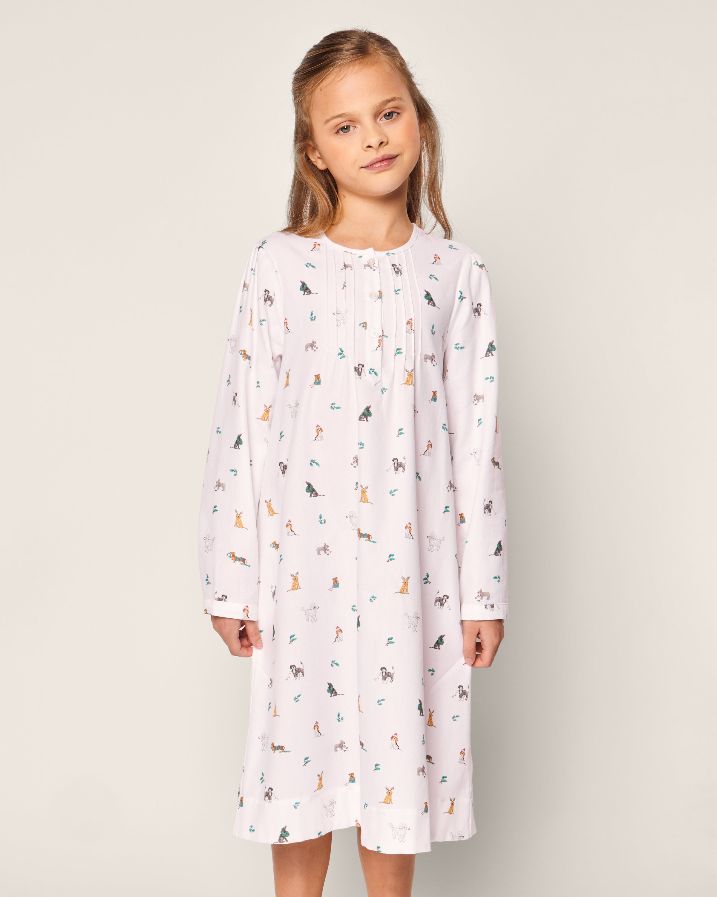A young girl in Petite Plumes Girls Twill Beatrice Nightgown in Jingle Paws stands against a plain backdrop. Her light brown hair frames her slight smile as she looks into the camera, exuding comfort and charm.