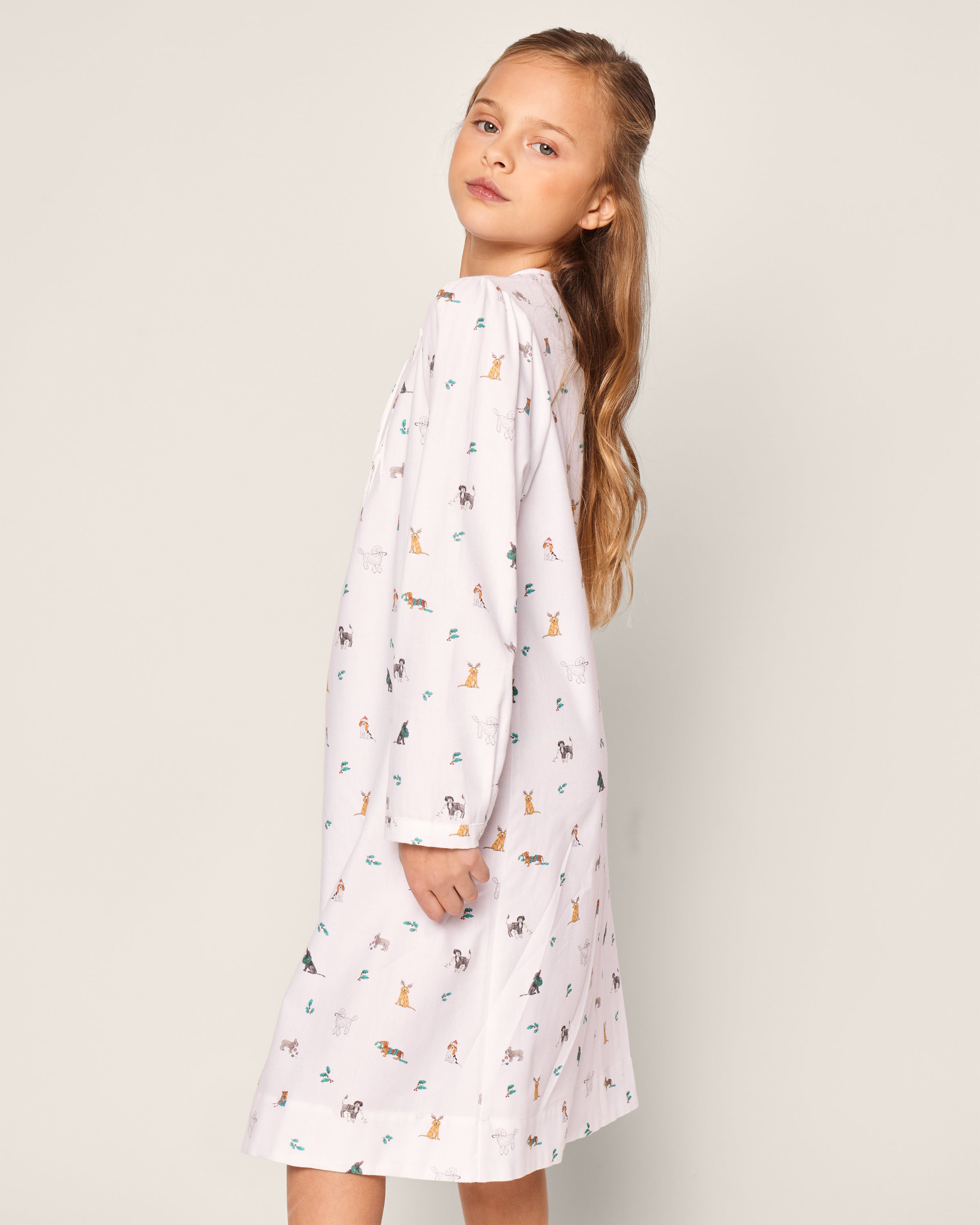 A young girl with long hair wears Petite Plumes Girls Twill Beatrice Nightgown in Jingle Paws, a white dress with colorful animal prints on chemical-free flame-retardant fabric. She stands sideways, slightly turning her head toward the camera, against a plain light-colored background.