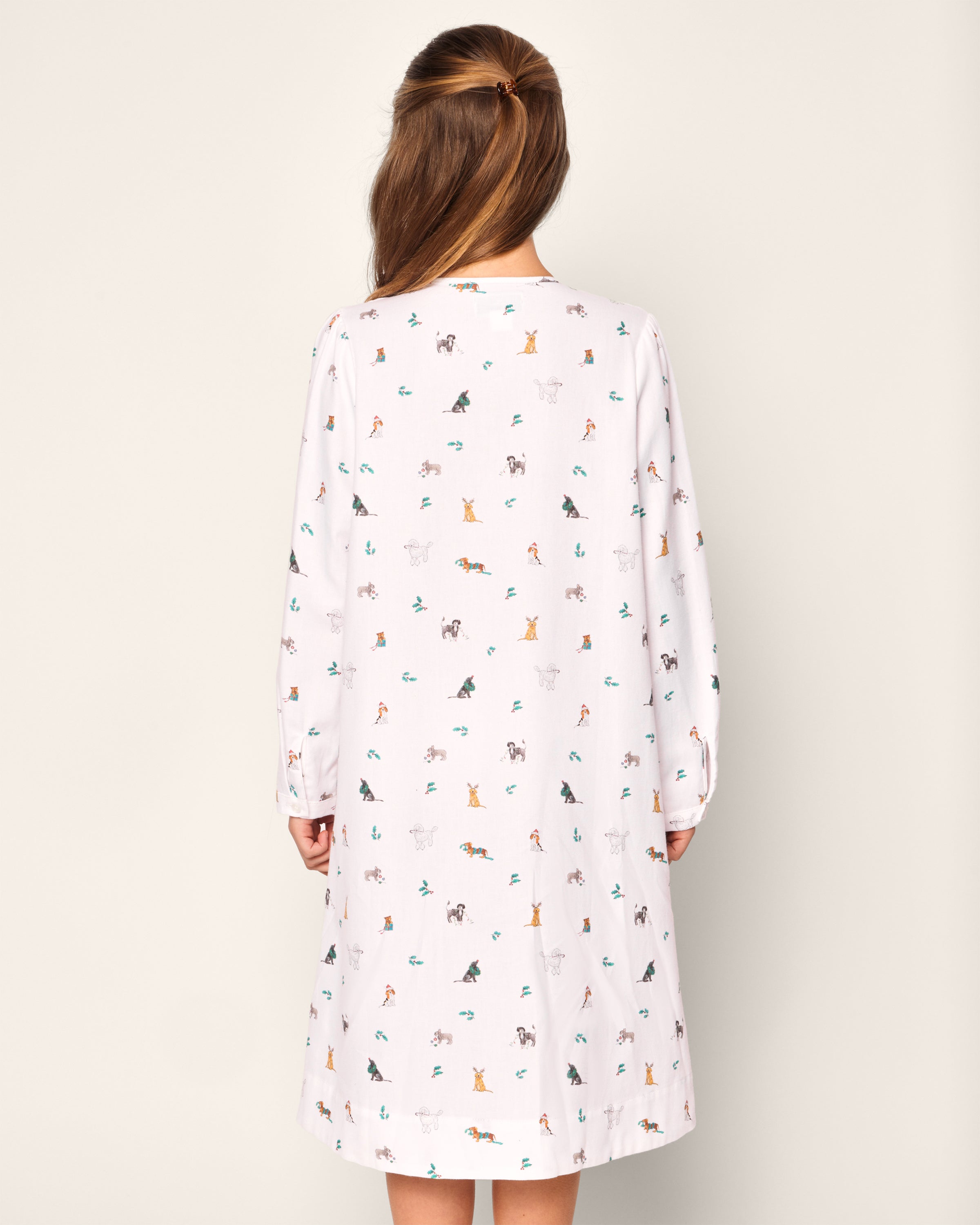 A person with long hair faces away, wearing Petite Plumes Girls Twill Beatrice Nightgown in Jingle Paws, a knee-length dress featuring colorful animal illustrations. Its crafted from chemical-free flame-retardant fabric for style and safety against a plain white backdrop.