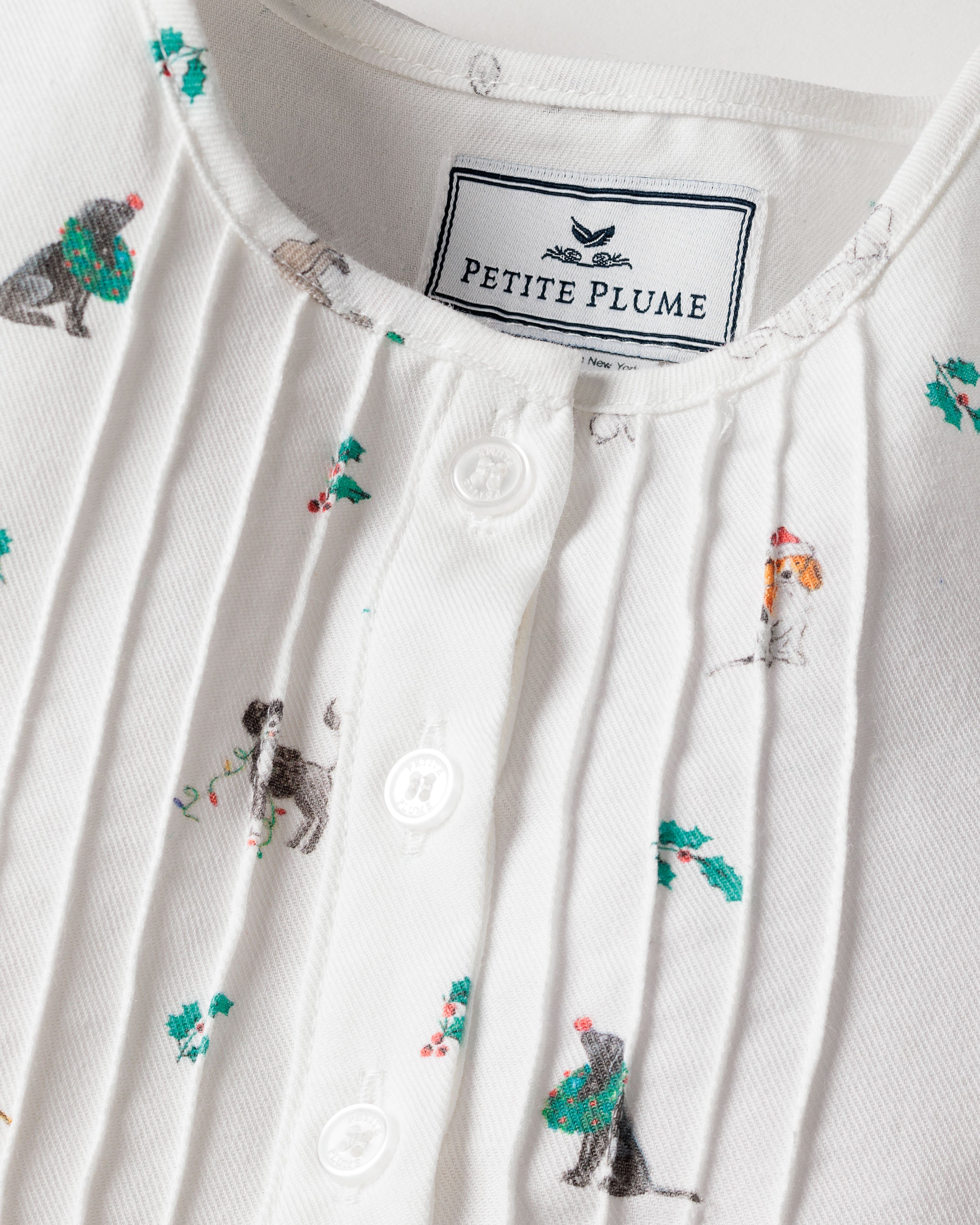 Close-up of Petite Plumes Girls Twill Beatrice Nightgown in Jingle Paws: a white childrens sleepwear shirt with dog illustrations in Santa hats, wreaths, and stockings. Made from chemical-free fabric, it features pleats and white buttons with a brand label near the collar for comfort and style.