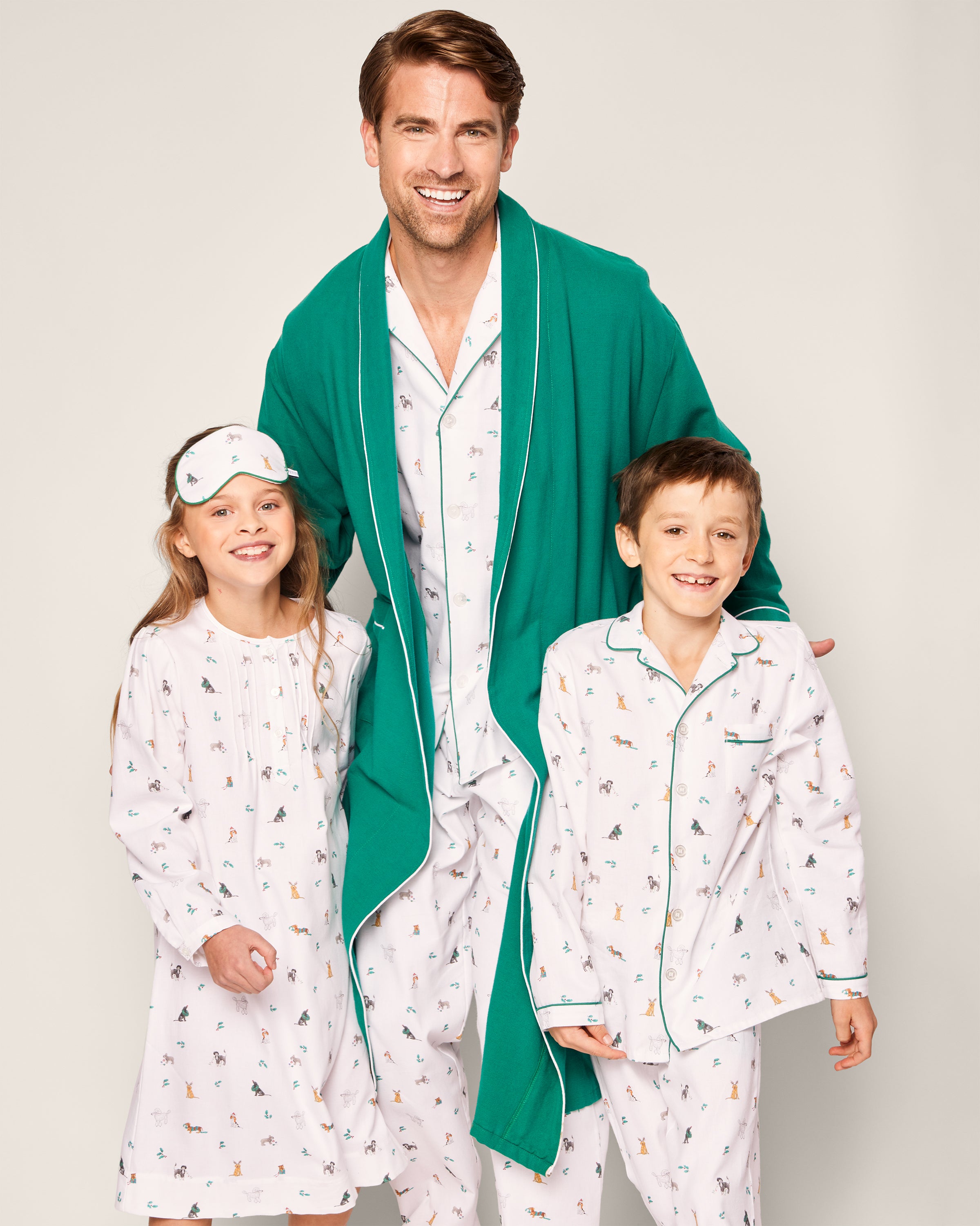 A man wearing a green robe and patterned pajamas stands with two smiling children in matching Petite Plume sleepwear. The girl wears the Girls Twill Beatrice Nightgown in Jingle Paws and a sleep mask, while her brother sports pajama pants made from chemical-free flame-retardant fabric.