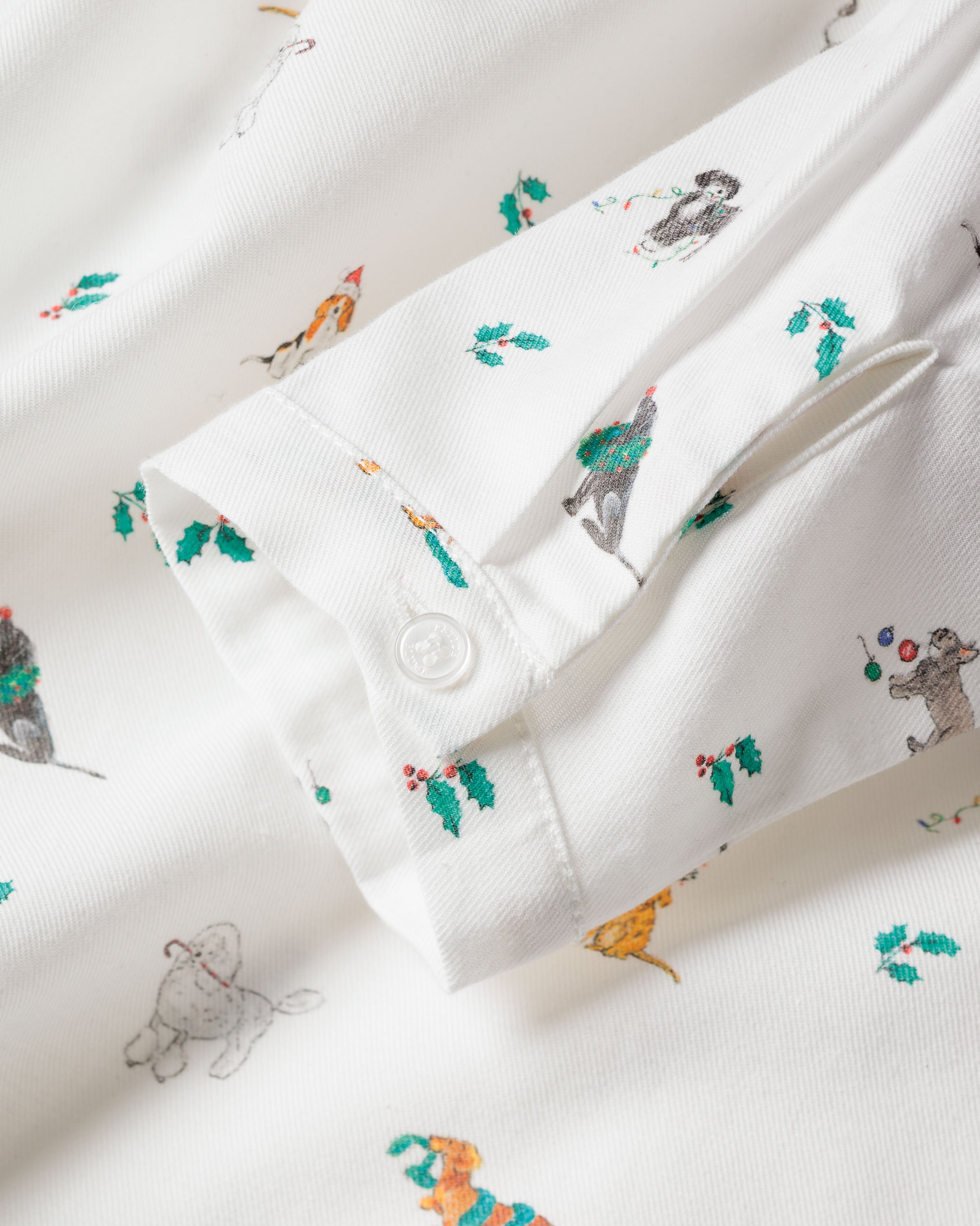 Close-up of Petite Plumes Girls Twill Beatrice Nightgown in Jingle Paws, showcasing a white fabric with festive animal illustrations and holly leaves. Ideal for childrens luxurious sleepwear, it features a buttoned placket with one button fastened and another nearby.