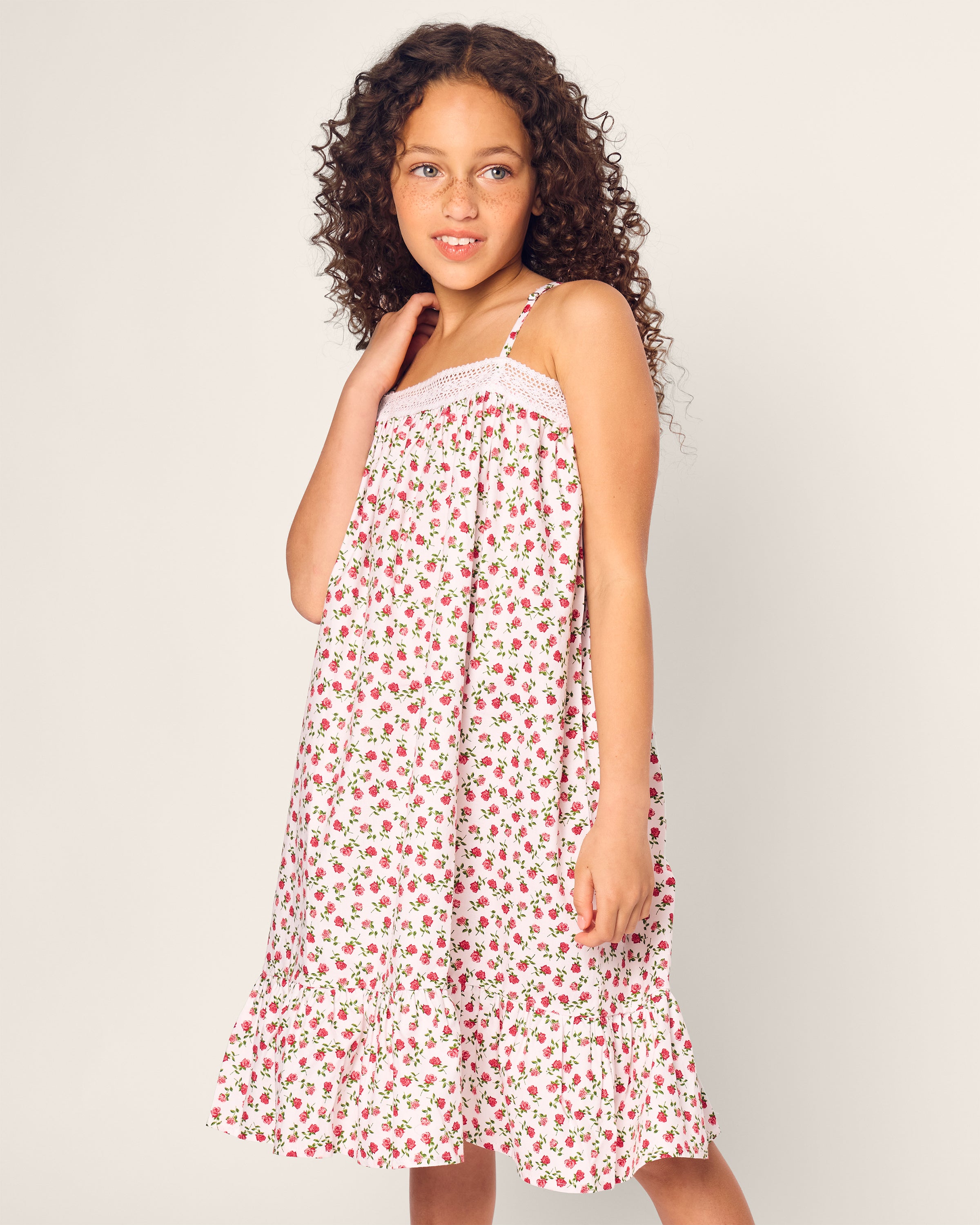 A young girl wearing Petite Plumes Girls Lily Nightgown in Petite Petals, made from soft, yarn-dyed cotton, stands against a plain background. Her long curly hair cascades over the white nightgown adorned with red floral patterns as she gazes to the side with a gentle smile.