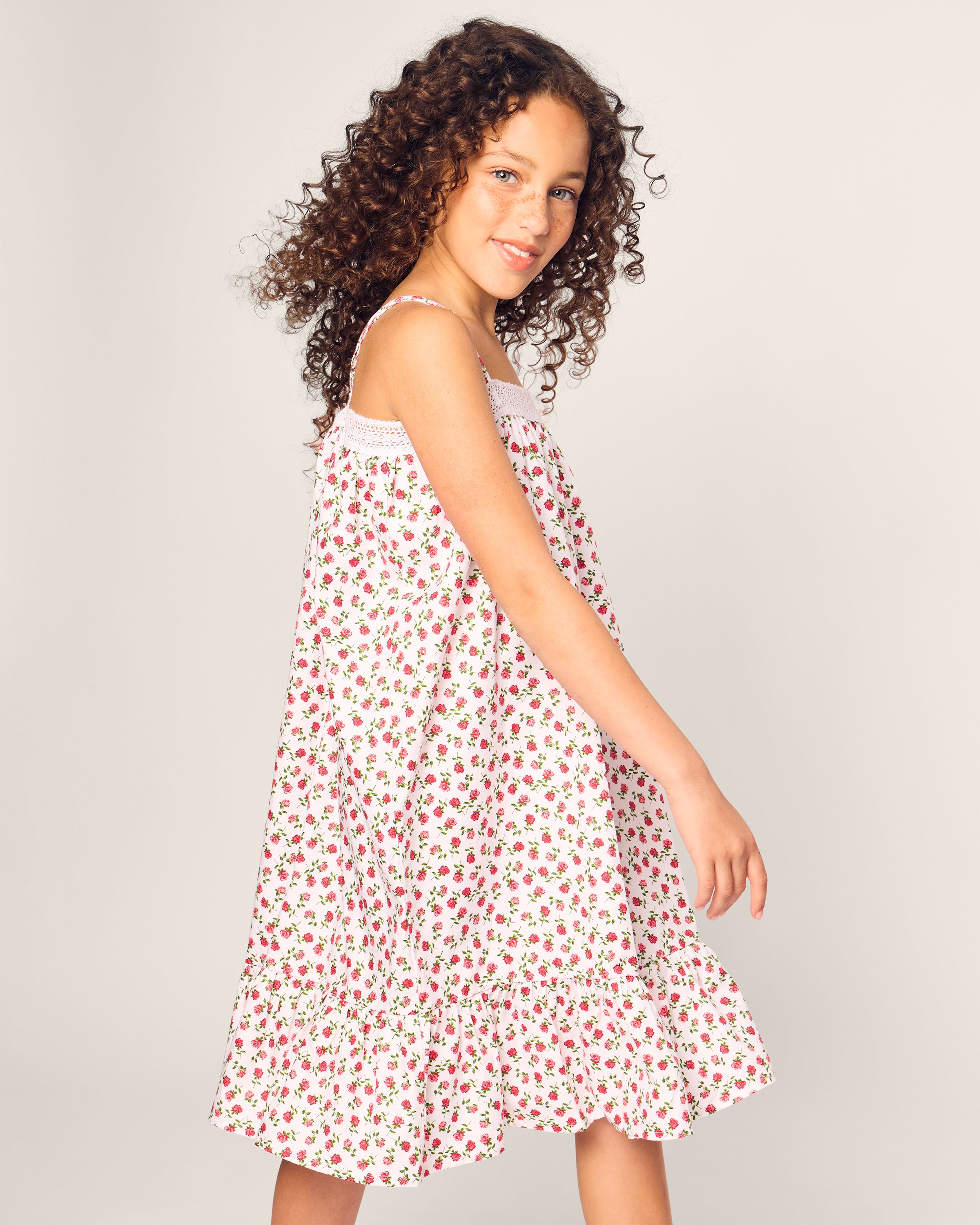 A young girl with curly hair smiles and poses in the Girls Lily Nightgown in Petite Petals by Petite Plume, featuring a red floral pattern. She stands against a plain light gray background, glancing over her shoulder.