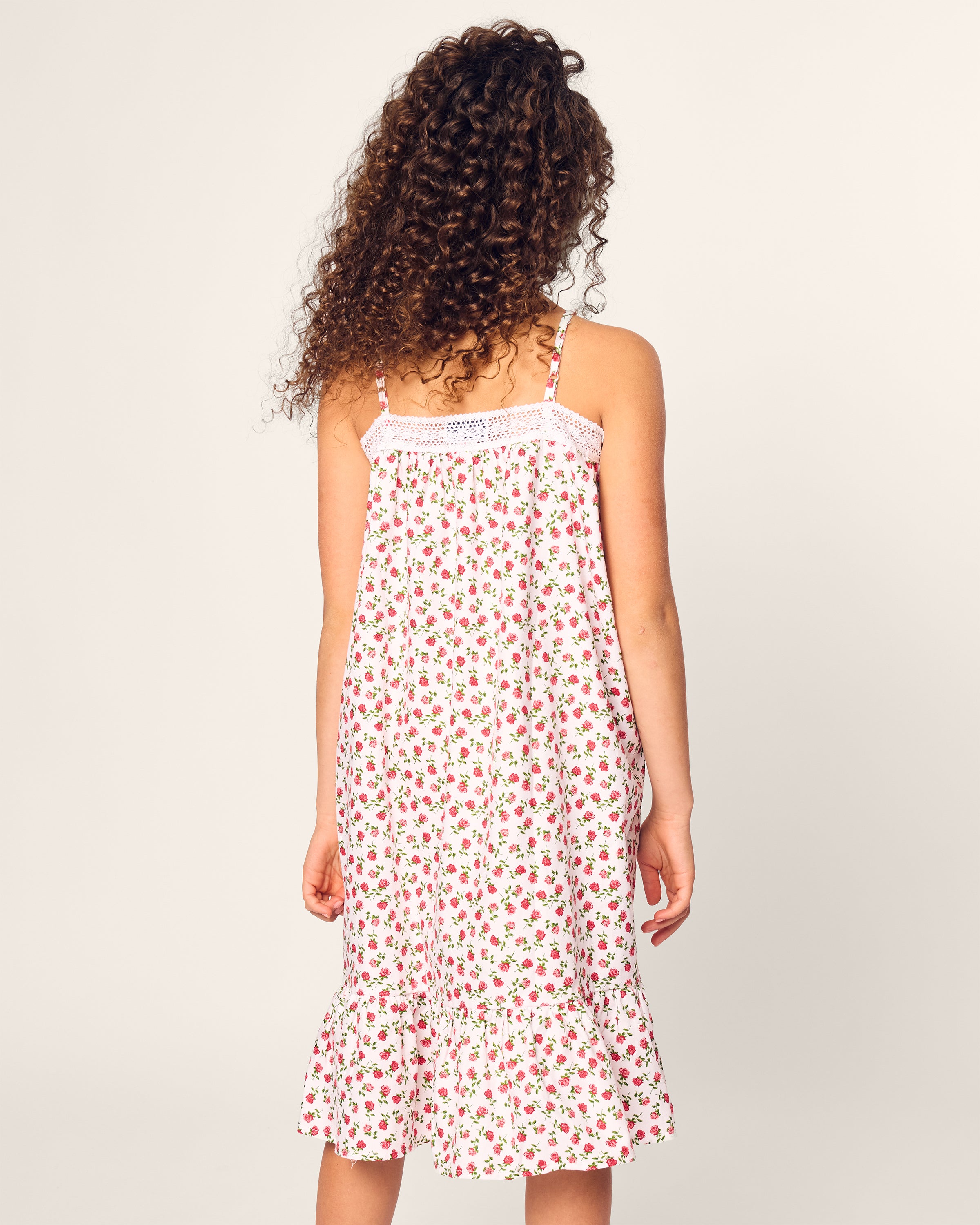 A curly-haired person in a sleeveless, knee-length white dress with a red floral pattern reminiscent of luxurious sleepwear stands against a plain background, facing away from the camera, evoking the elegance of Petite Plumes Girls Lily Nightgown in Petite Petals.